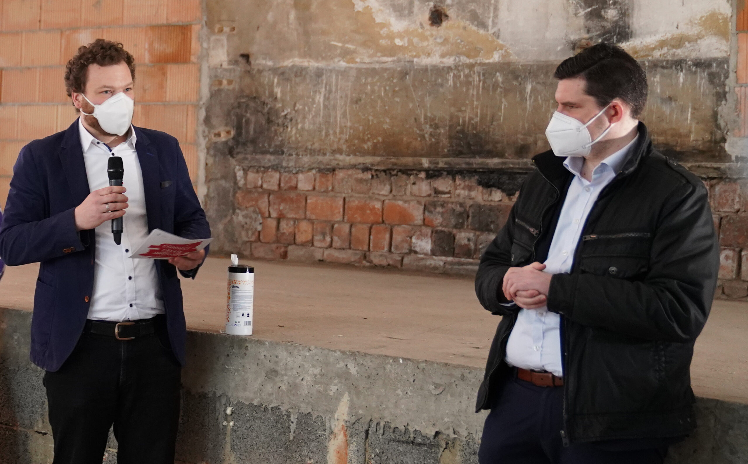 Zwei Männer mit Masken stehen im Gebäude der Hochschule Coburg. Einer hält ein Mikrofon und ein Blatt Papier, während der andere mit gefalteten Händen zuhört. In dem Raum mit seinen Ziegel- und Betonwänden steht auf einem Sims eine Flasche Desinfektionsmittel, was das Engagement der Hochschule für Sicherheit unterstreicht.