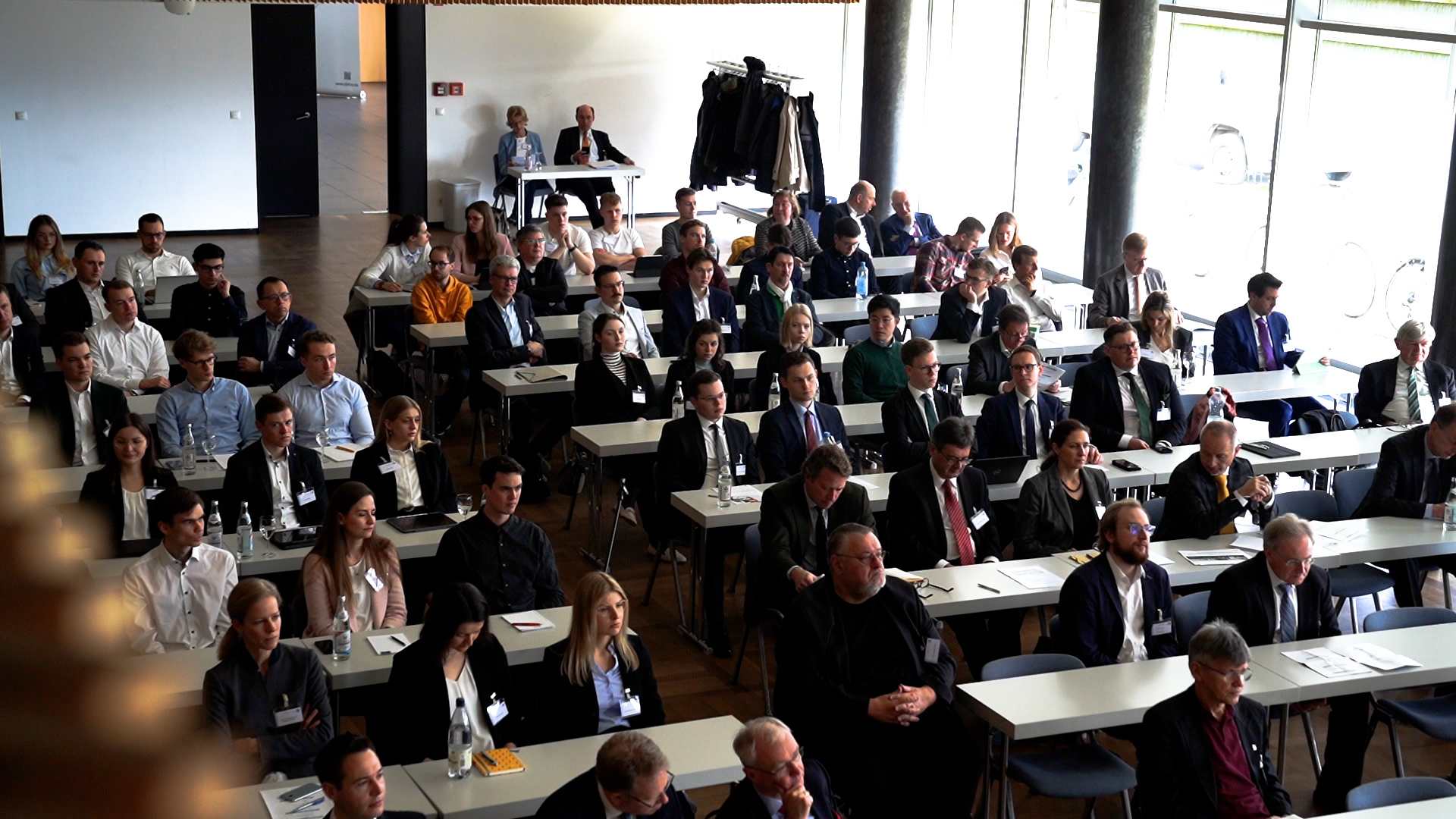 Eine große Gruppe von Menschen in formeller Kleidung sitzt in Reihen bei einer Konferenz oder einem Seminar der Hochschule Coburg. Sie blicken nach vorne und hören einem Redner in der ersten Reihe zu. Der Raum ist gut beleuchtet, natürliches Licht fällt durch die Seitenfenster.