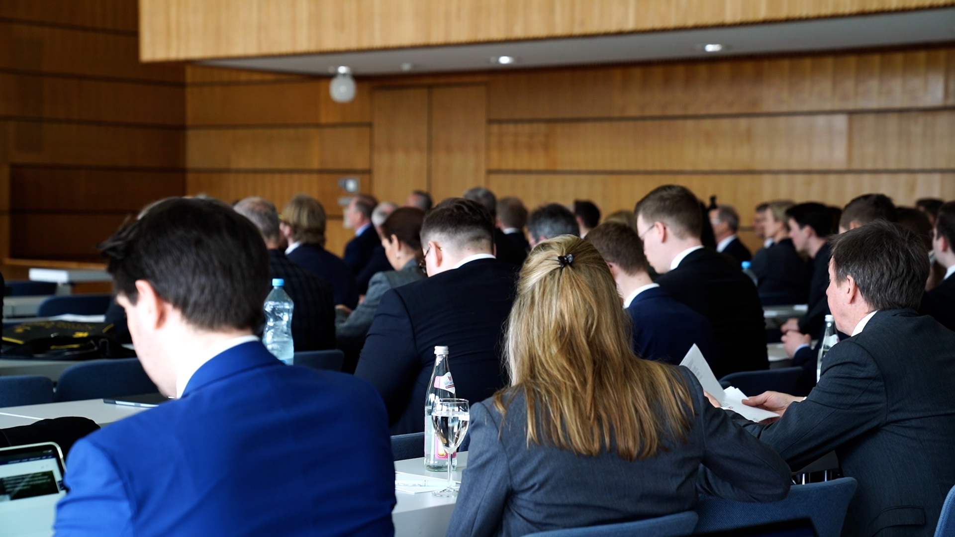 Zuhörer in formeller Kleidung sitzen bei einer Konferenz der Hochschule Coburg einem Redner gegenüber. Der Raum ist mit Holz verkleidet und hell beleuchtet. Einige Teilnehmer machen sich Notizen, während andere auf einen Bildschirm schauen. Auf den Tischen stehen Wasserflaschen.