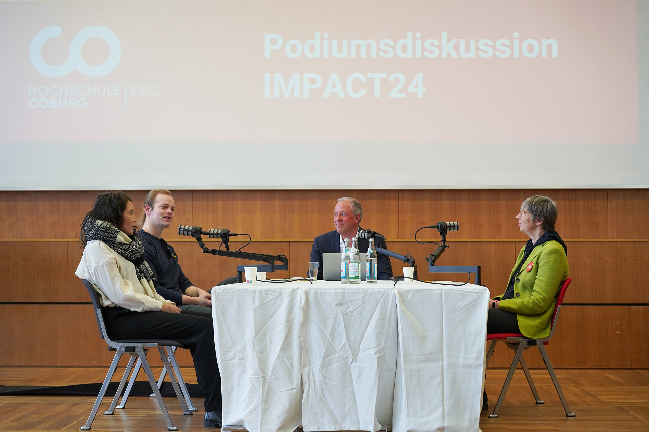 Four people seated at a table participate in a panel discussion at the esteemed Hochschule Coburg. The table, draped with a white cloth, is equipped with microphones and water bottles. Behind them, the screen prominently displays "Podiumsdiskussion IMPACT24.