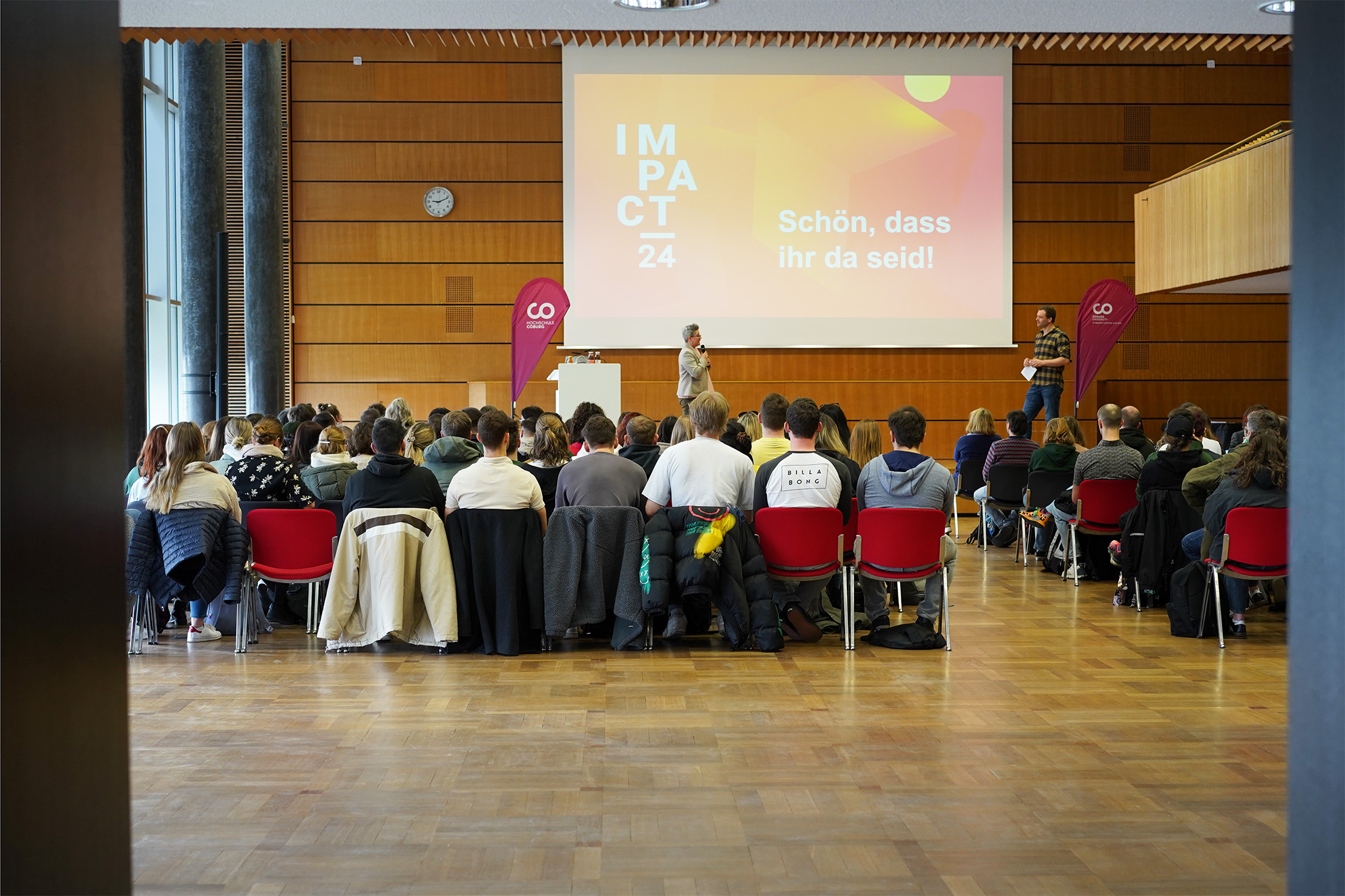 A group of people seated in a conference room at Hochschule Coburg faces a presentation screen displaying "IMPACT 24" and "Schön, dass ihr da seid!" Two speakers stand on stage, amidst wooden paneling and red chairs.