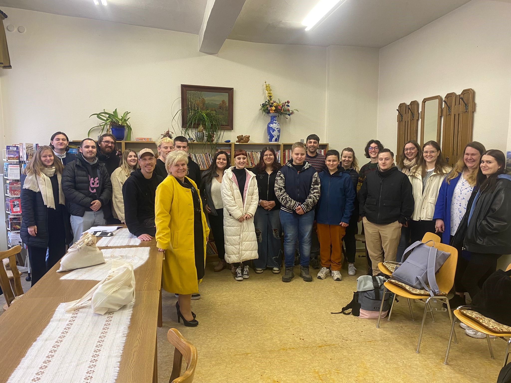 Eine Gruppe von Menschen lächelt für ein Foto in einem Raum der Hochschule Coburg mit einem langen Tisch und mehreren Stühlen. Eine Frau in einem gelben Mantel steht vorne. Der Raum ist mit Pflanzen, einem Gemälde und Holzdekor an den Wänden geschmückt. Auf dem Tisch und den Stühlen liegen verstreut einige Taschen.