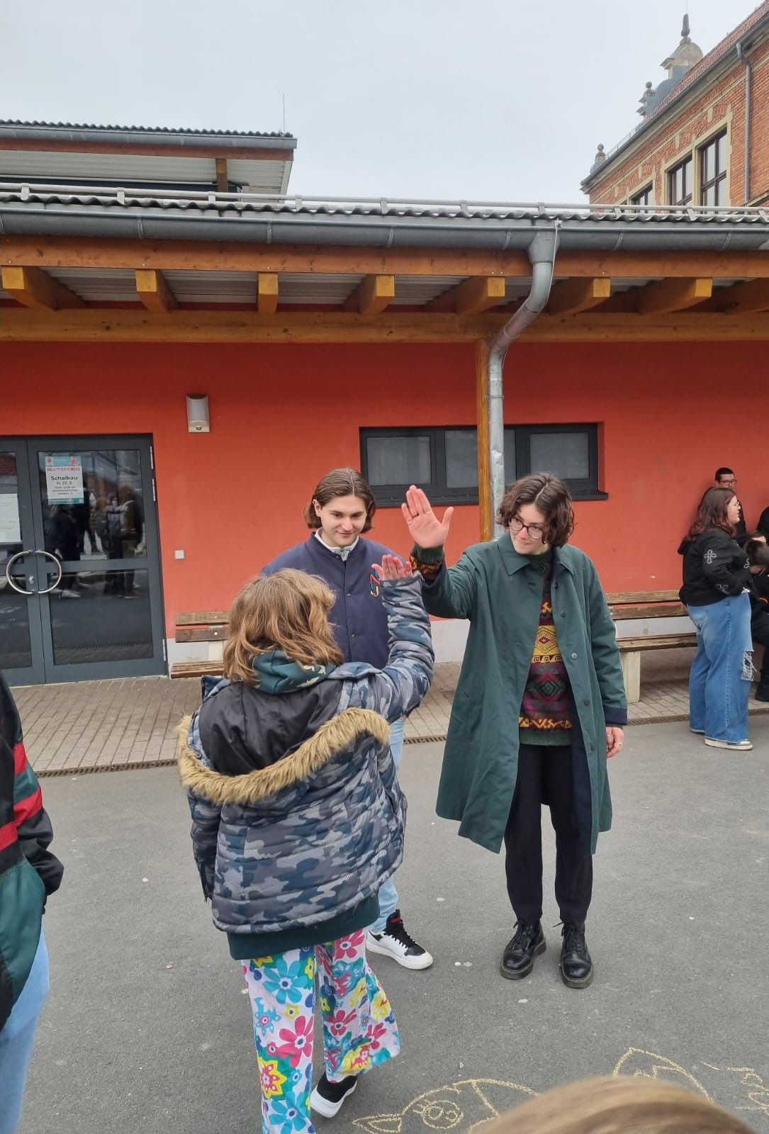 A person wearing a long coat and glasses gives a high five to someone in a colorful outfit outside Hochschule Coburg. Another person stands nearby, witnessing the cheerful moment in front of an orange building on a cloudy day.