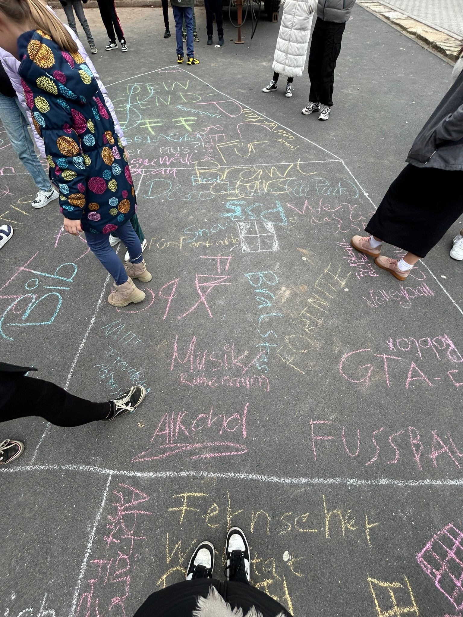 People gather around colorful chalk drawings on a gray pavement, featuring words like "Musik," "Fernseher," and "Fussball." Among the visible feet, Hochschule Coburg students appear to be engaged in a lively group activity or game.