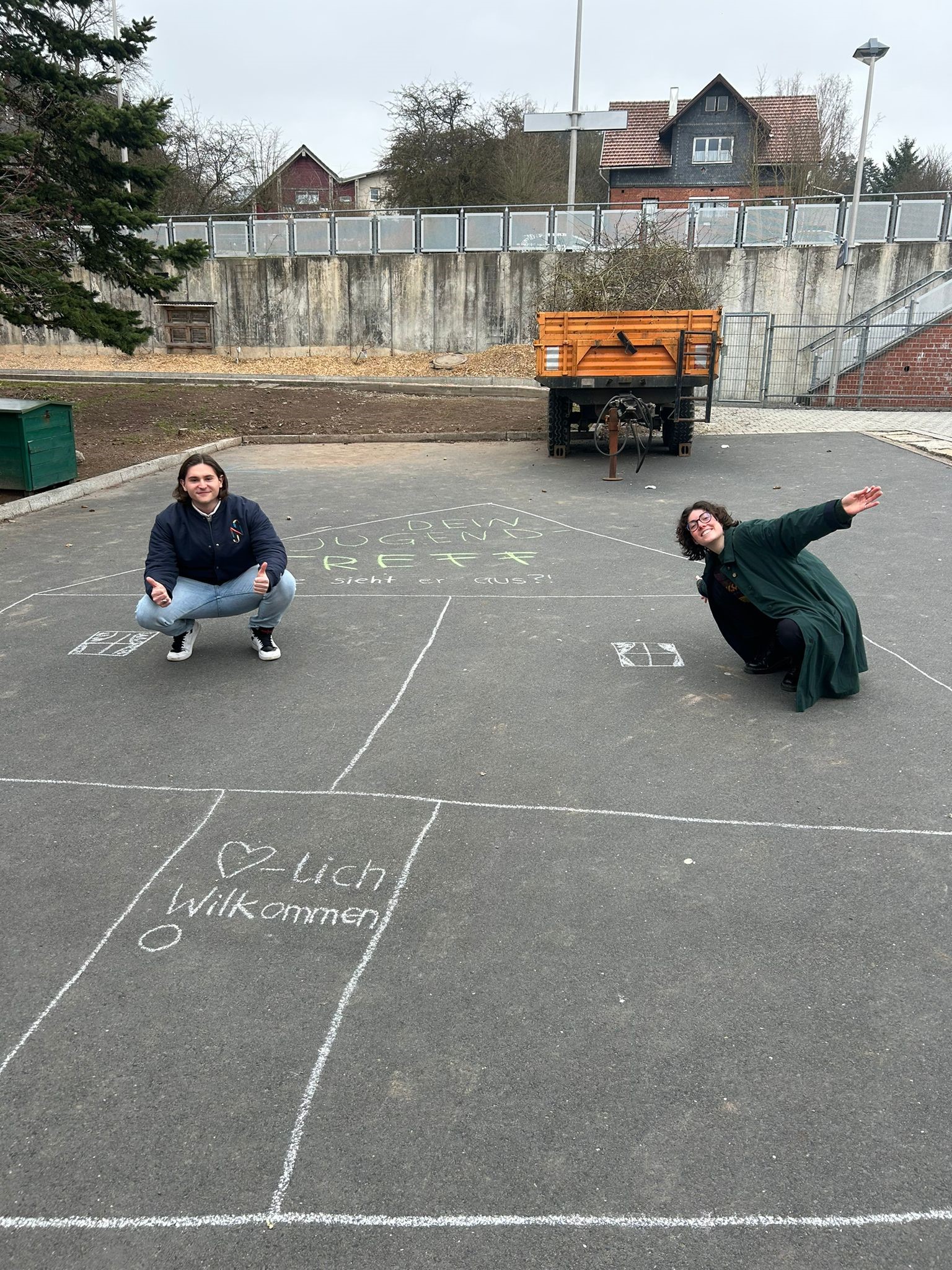 Two people happily pose on a street adorned with chalk drawings near Hochschule Coburg. One kneels while the other crouches nearby. Chalk writings include "Herzlich Willkommen" with a heart, and in the background, there's a fenced area with trees and a truck.