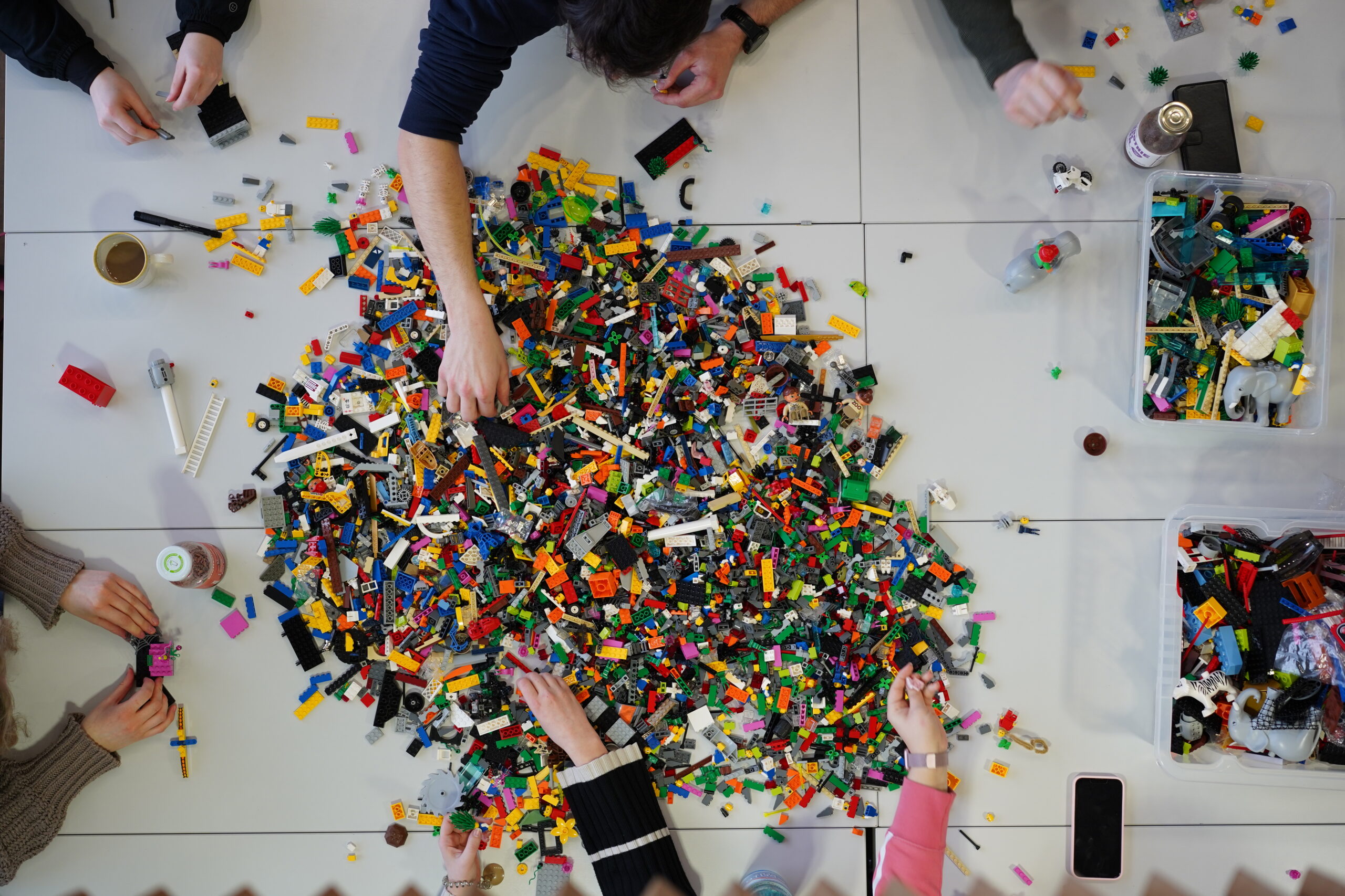 A group of people from Hochschule Coburg is seated around a table filled with colorful LEGO pieces, each reaching in to select pieces from the central pile, with drinks and containers adding to the lively atmosphere.