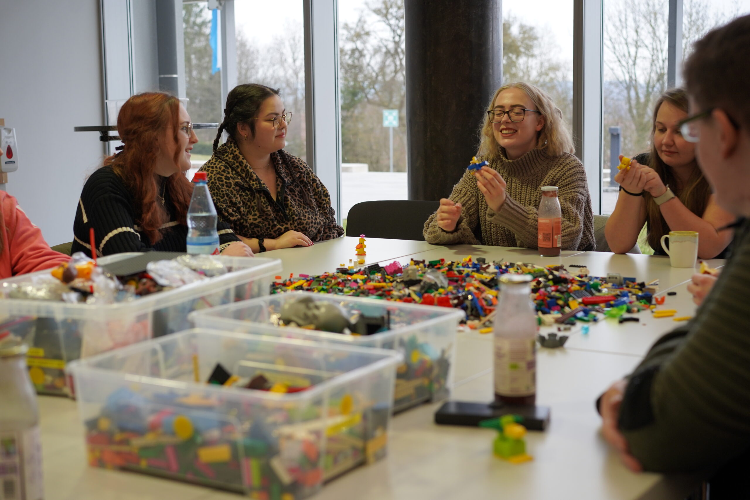 A group of people from Hochschule Coburg sit around a table covered with LEGO bricks, engaged in conversation. Some hold LEGO pieces while smiling. Bottles and drinks are also on the table. Large windows in the background offer a view of trees.