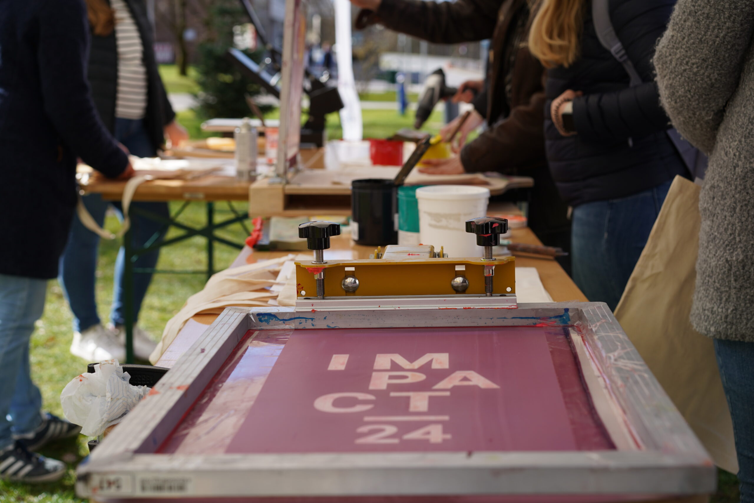 An einem langen Tisch im Freien drucken Mitarbeiter der Hochschule Coburg im Siebdruckverfahren. Im Mittelpunkt steht ein Siebdruck, auf dem „IMPACT 24“ teilweise zu sehen ist. Verschiedene Druckwerkzeuge und Farben liegen verstreut herum, während die Teilnehmer mit Begeisterung ihrer kreativen Tätigkeit nachgehen.