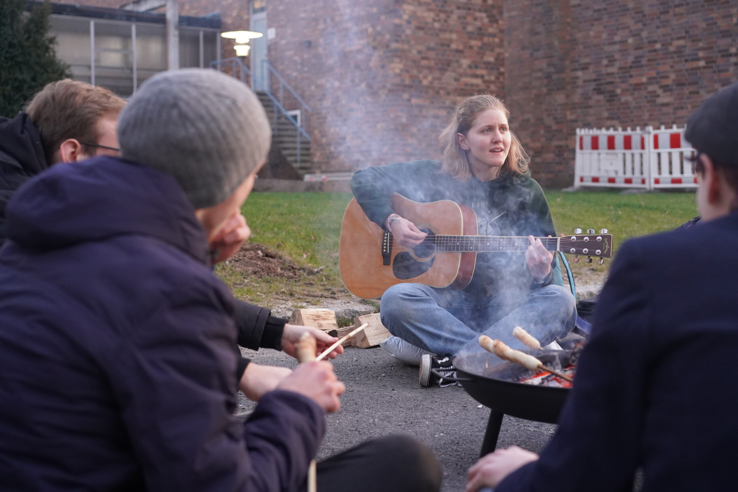 A group sits around a small campfire outside Hochschule Coburg, with one person playing an acoustic guitar and singing. Others hold sticks with bread dough over the fire, enjoying an outdoor, casual gathering near the brick building.