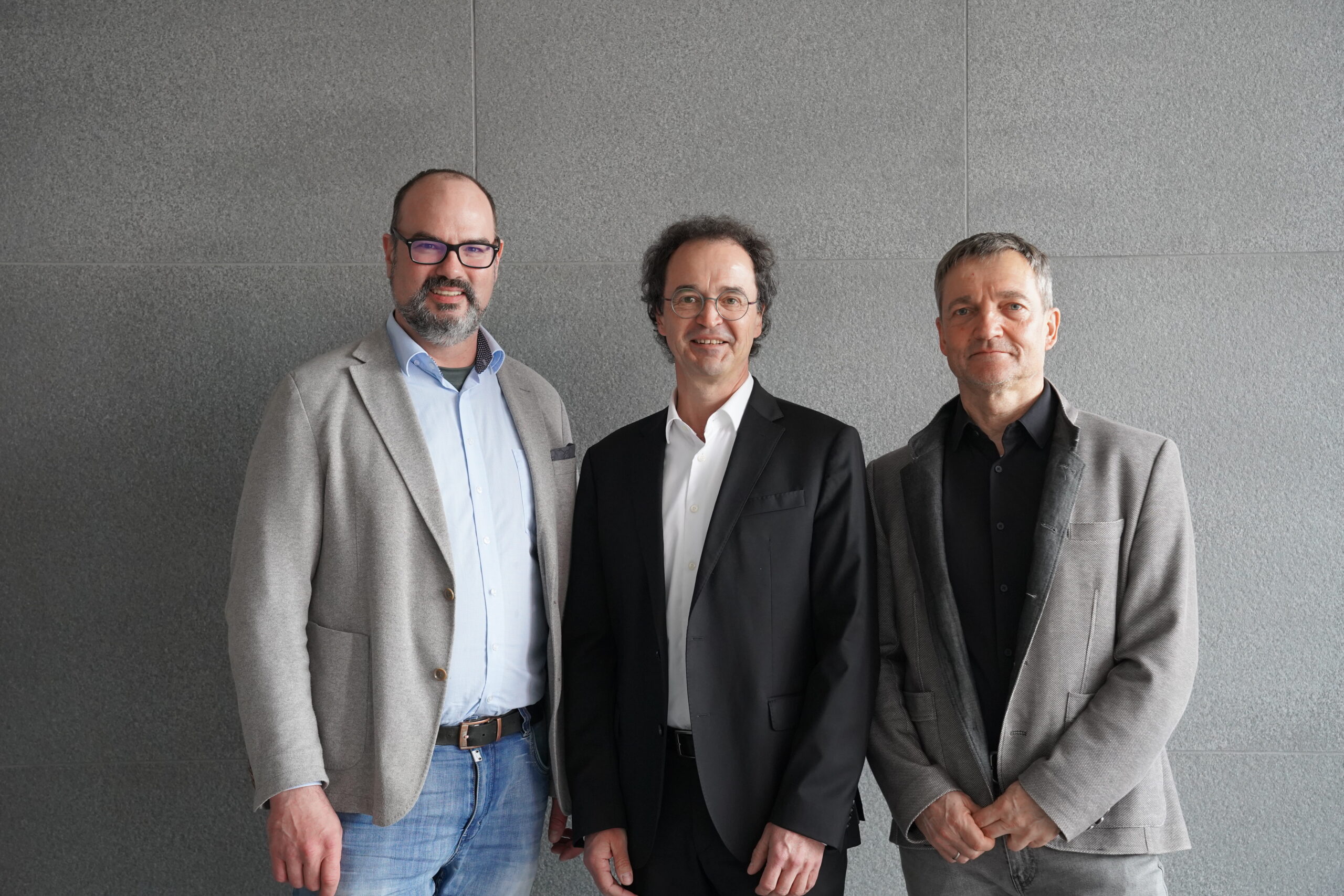 Three men are standing side by side, all wearing blazers. The man on the left, possibly a Hochschule Coburg alumnus, sports glasses and a light gray blazer with jeans. The man in the middle dons a black suit, while the one on the right looks sharp in a gray blazer and darker shirt. They all smile slightly.