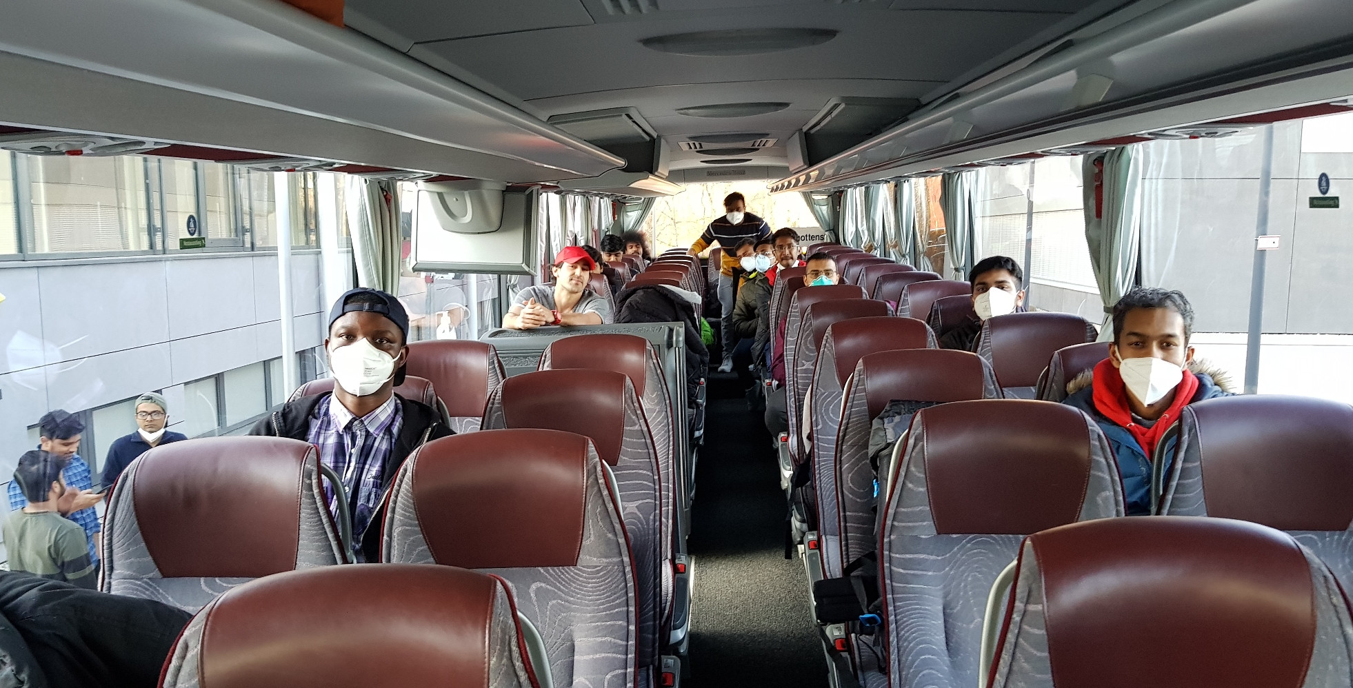 Eine Gruppe von Menschen, einige davon mit Masken, sitzt mit Abstand in einem leeren Shuttlebus der Hochschule Coburg. Der Innenraum ist modern, mit kastanienbraunen und grauen Sitzen. Sonnenlicht fällt durch die Fenster herein.