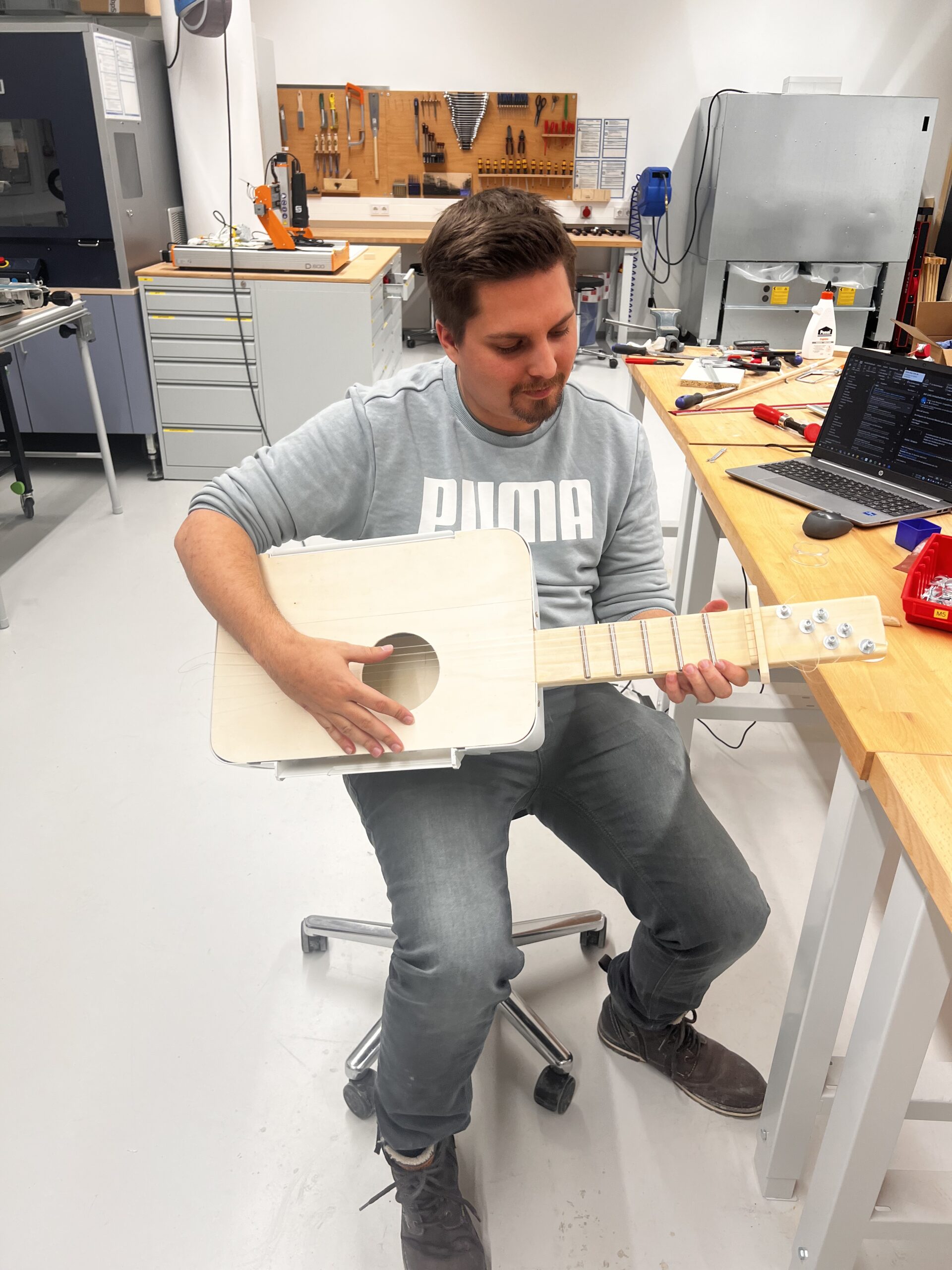 In einer gemütlichen Werkstatt der Hochschule Coburg sitzt eine Person in einem grauen Sweatshirt auf einem Hocker und spielt gekonnt ein hölzernes gitarrenähnliches Instrument. Um sie herum stehen Werkbänke, Werkzeuge und Geräte, die die kreative Atmosphäre des Raumes widerspiegeln.