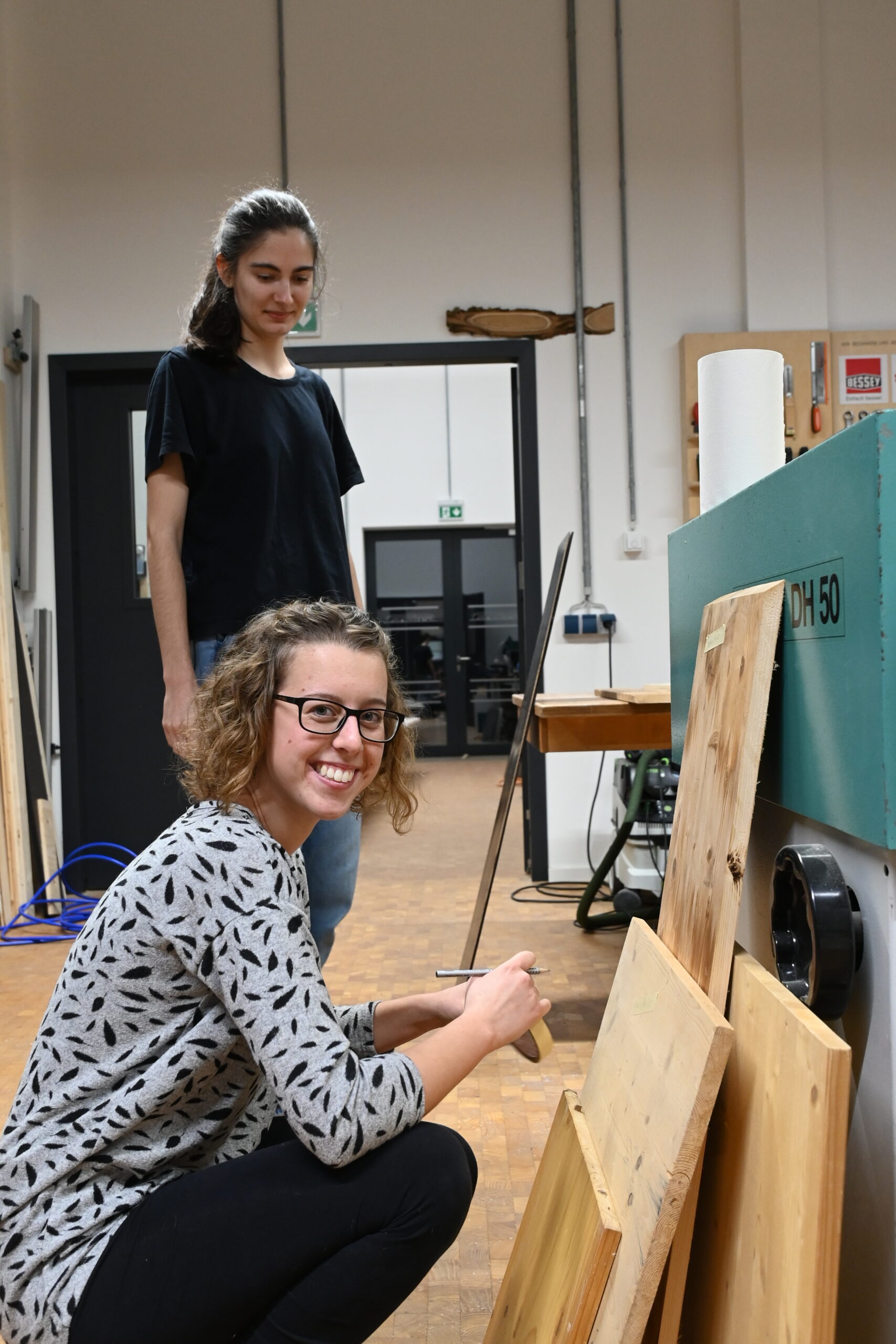 Zwei Frauen vertiefen sich in ihr Holzhandwerk an der Hochschule Coburg. Die eine hockt lächelnd und hantiert für die Kamera mit Holzbrettern, die andere steht dahinter und beobachtet nachdenklich. Um sie herum sind sie von Geräten und Werkzeugen umgeben, die eine authentische Werkstattatmosphäre schaffen.