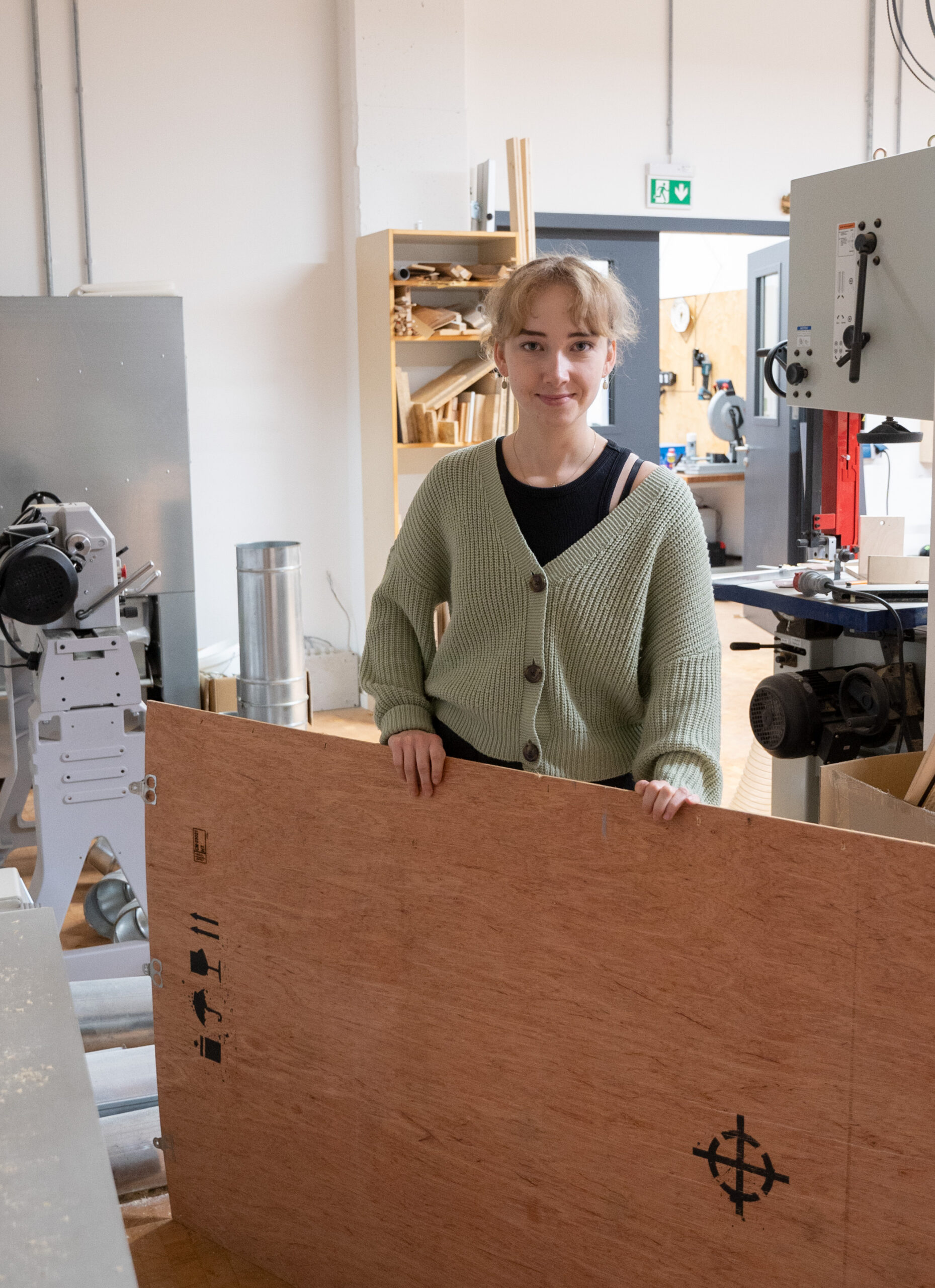 In einer Werkstatt voller Kreativität steht eine Person neben einem großen Holzbrett, trägt eine grüne Strickjacke und lächelt herzlich in die Kamera. Sie repräsentiert die Hochschule Coburg und ist von einer Reihe von Werkzeugen und Geräten umgeben, die den Geist des praktischen Lernens verkörpern.