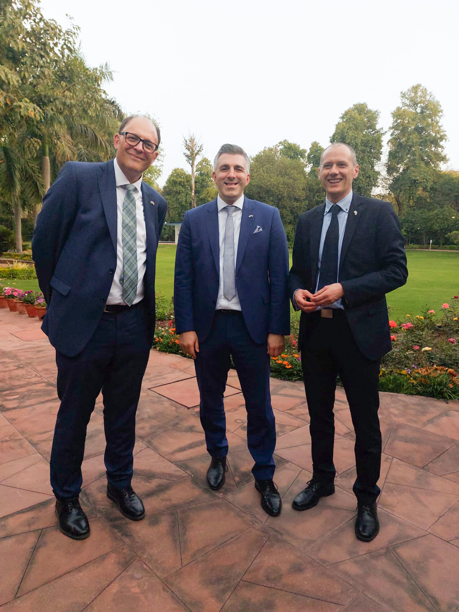 Three men in suits stand on a patio with a garden in the background at Hochschule Coburg. They are smiling and wearing ties, with trees and flowerbeds visible behind them.