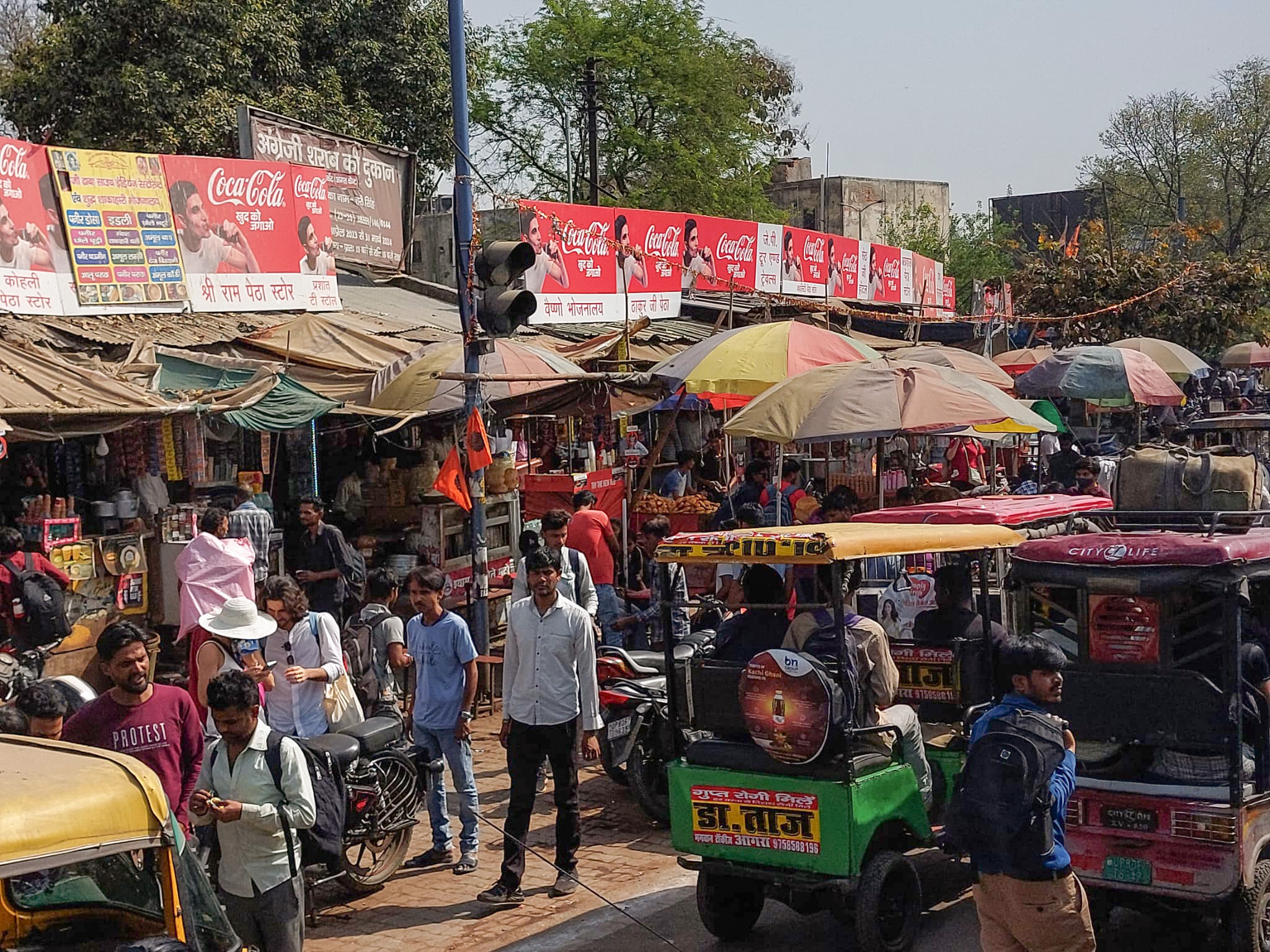 A bustling street market scene unfolds with colorful stalls selling various goods near Hochschule Coburg. People weave through motorbikes and auto rickshaws, while bright banners and signs above the stalls amplify the vibrant atmosphere.