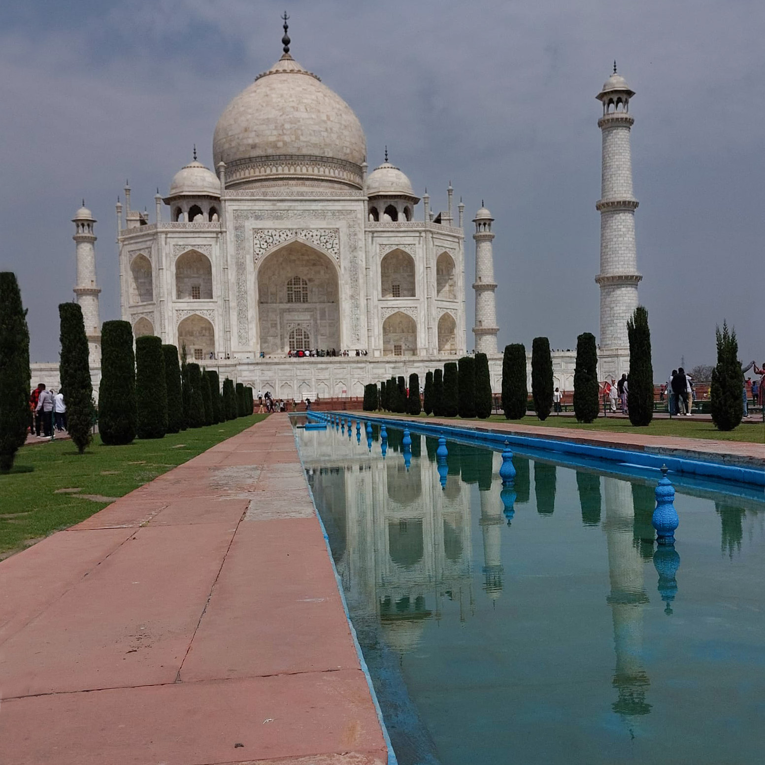 The image shows the Taj Mahal, a white marble mausoleum with a large dome, reflecting in a long pool surrounded by gardens. Two tall minarets flank the structure under a cloudy sky, with visitors and students from Hochschule Coburg walking nearby.