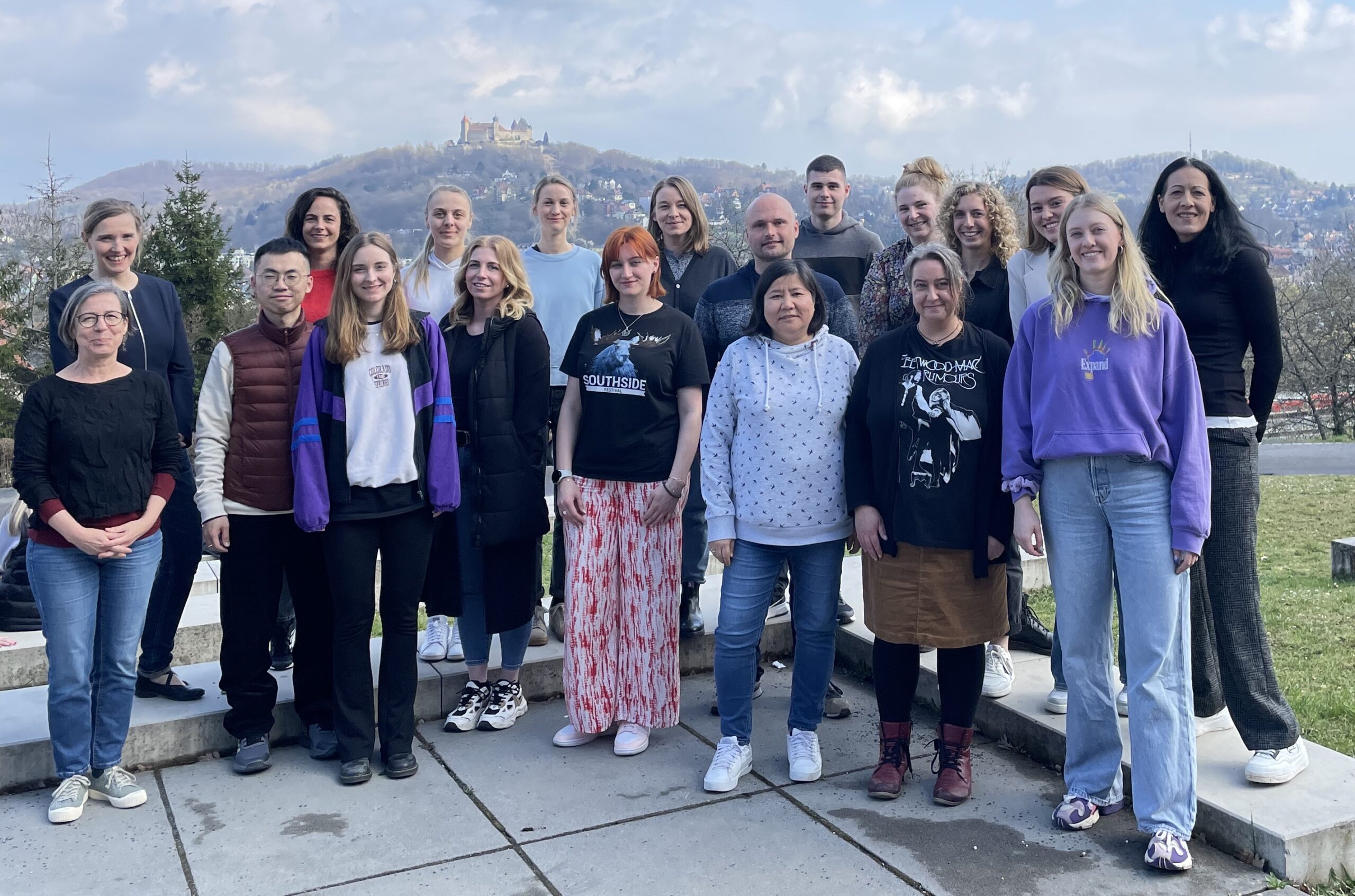 A group of twenty people, including students and staff from Hochschule Coburg, poses outdoors with a scenic view of a hilltop castle in the background. The men and women, dressed casually, stand smiling on a paved area surrounded by grass and trees.