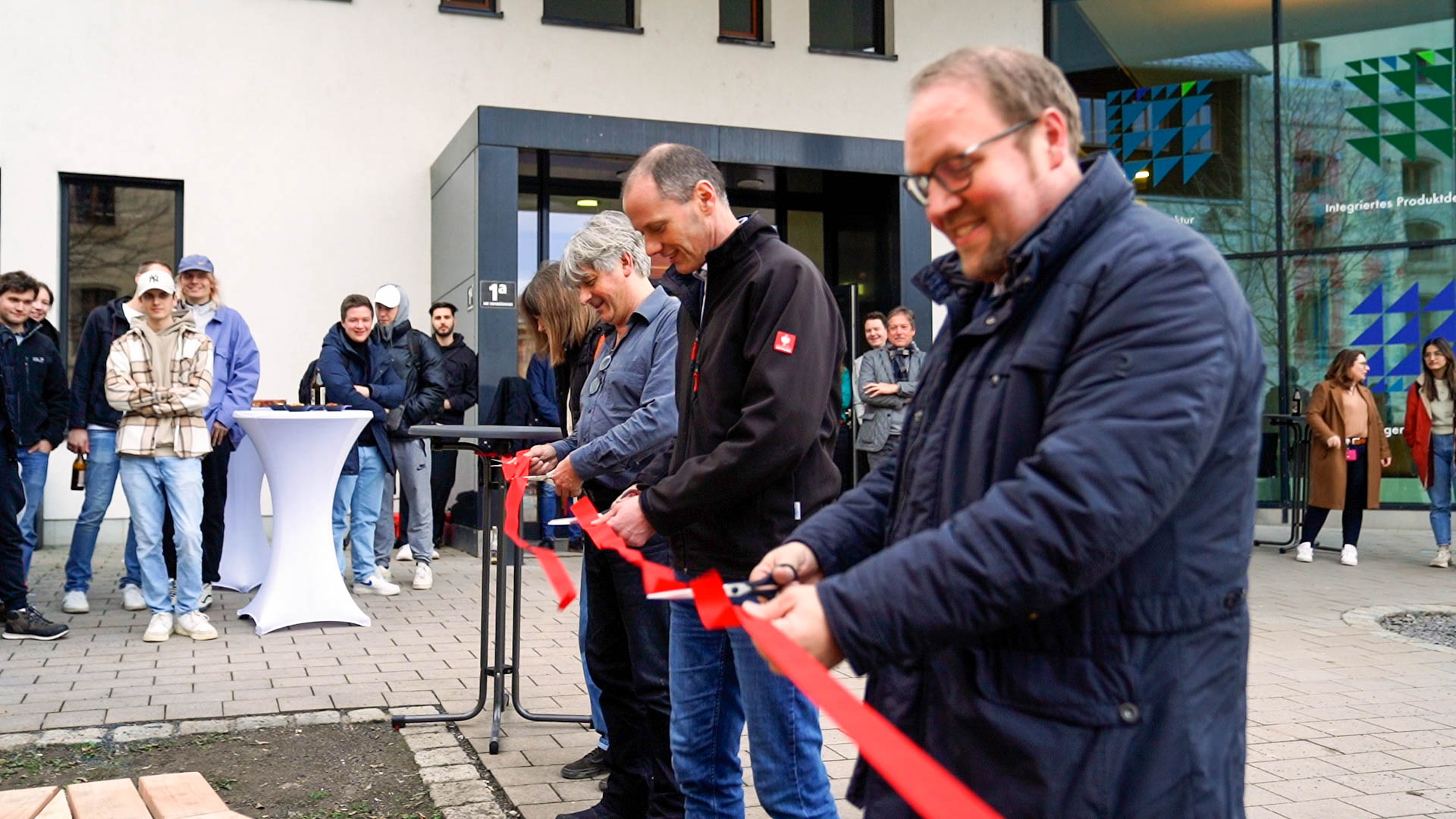 Eine Gruppe von Menschen steht stolz vor einem Gebäude der Hochschule Coburg und lächelt, während sie feierlich mit einer Schere ein rotes Band durchschneidet. Im Hintergrund haben sich Zuschauer versammelt, einige halten Kameras in den Händen, um den Moment festzuhalten. In der Nähe warten mit weißen Tischdecken bedeckte Tische auf ihre Gäste.