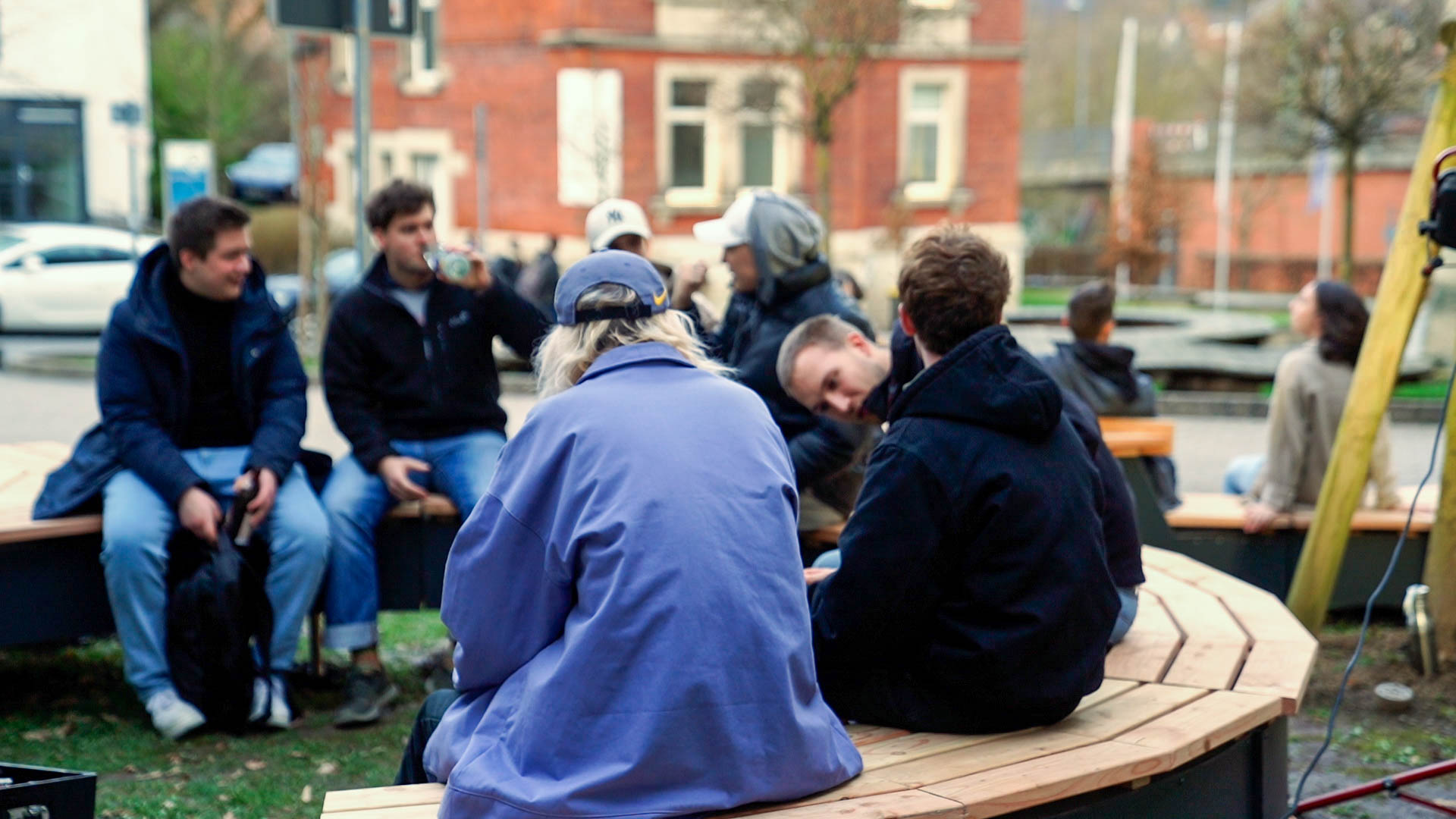 Eine Gruppe von Menschen sitzt auf geschwungenen Holzbänken im Freien vor der Hochschule Coburg und unterhält sich. Warm gekleidet unterhalten sie sich in einer lockeren und geselligen Umgebung mit Gebäuden, Bäumen und einem geparkten Auto im Hintergrund.