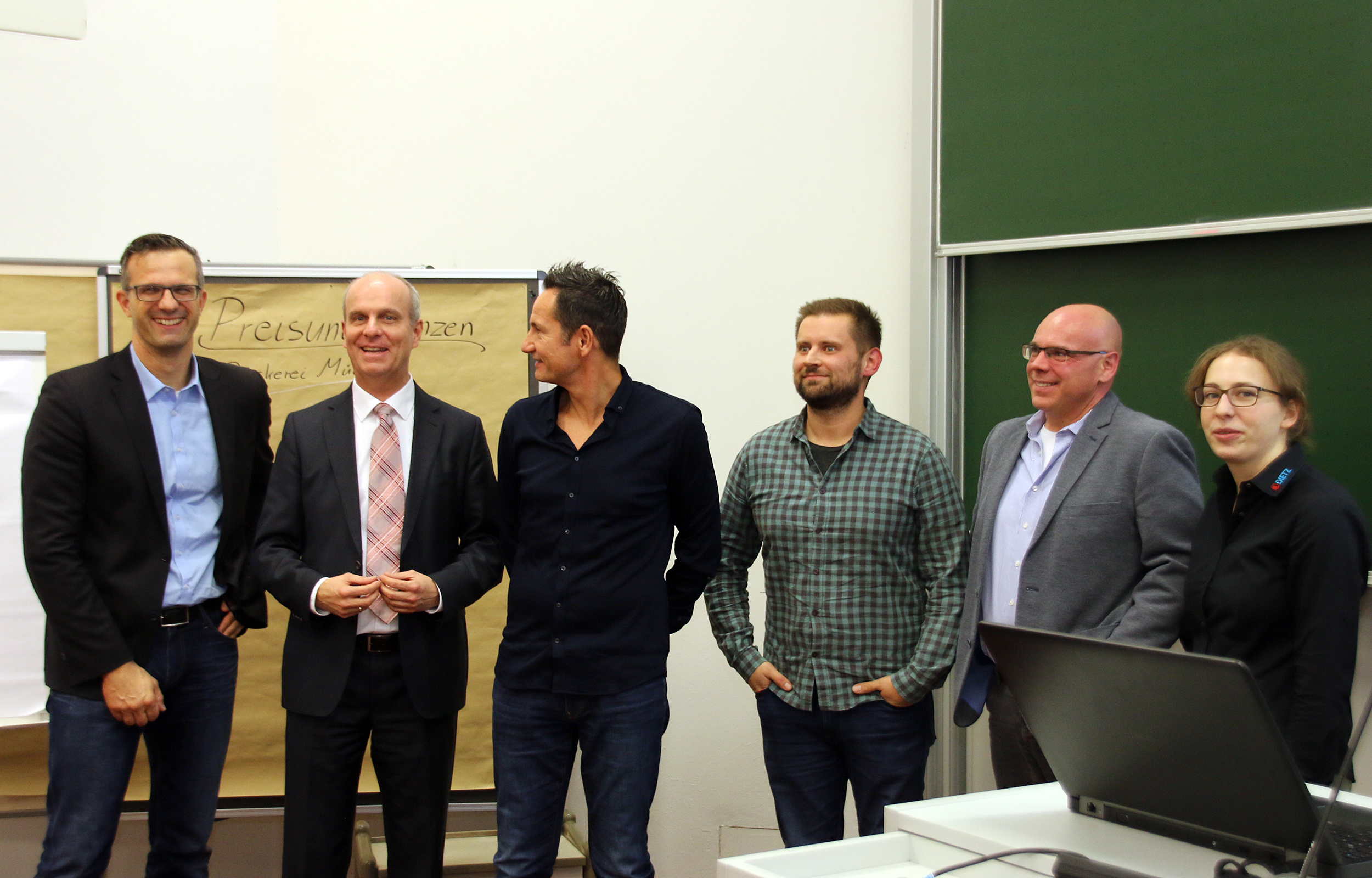 Eine Gruppe von sechs professionell gekleideten Personen steht in einem Unterrichtsraum der Hochschule Coburg. Einige lächeln. Auf einem Tisch steht ein Laptop, im Hintergrund ein Flipchart und eine große Tafel.