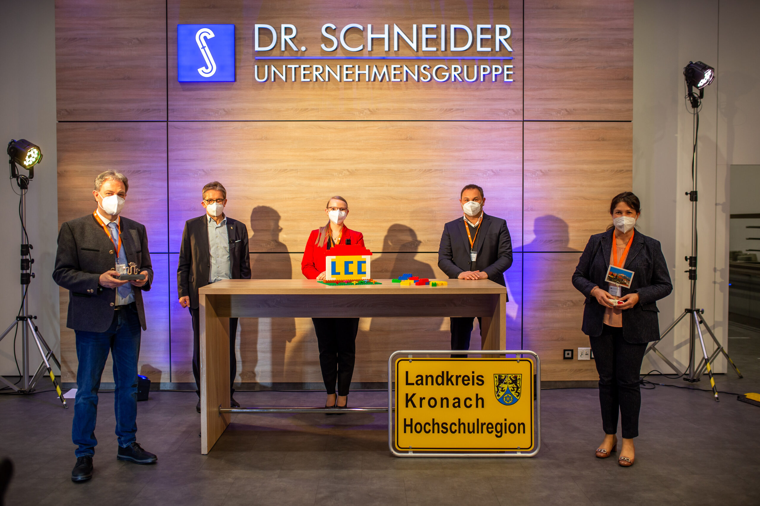 Fünf Menschen mit Masken stehen in einem Raum mit der Aufschrift „Dr. Schneider Unternehmensgruppe“. Sie halten Gegenstände in den Händen, und die Schilder „Landkreis Kronach HochSchulregion“ und „Hochschule Coburg“ sind gut sichtbar angebracht. Ein Holztisch und helle Beleuchtung sorgen für eine formelle Atmosphäre.