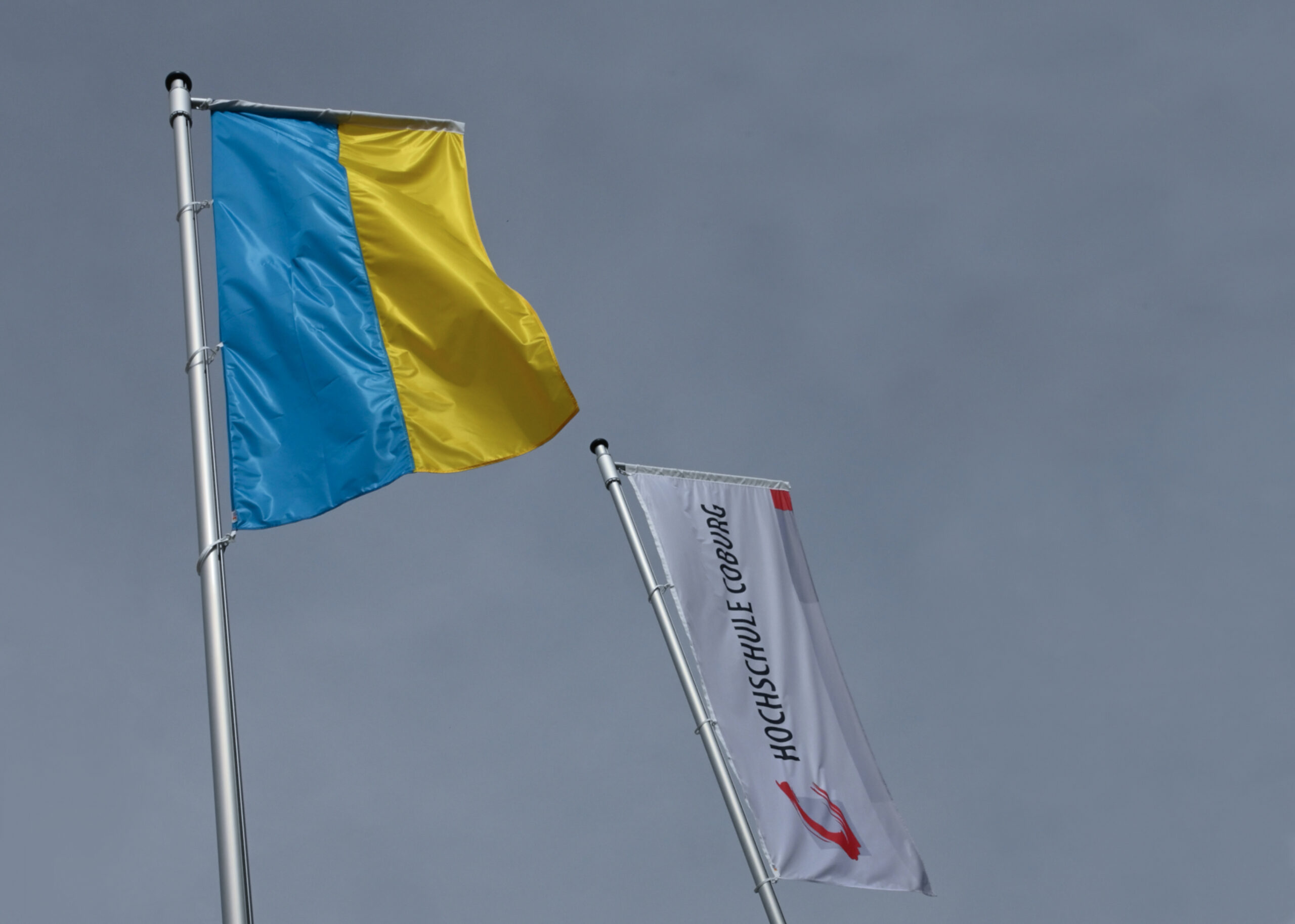 Zwei Flaggen an Fahnenmasten vor einem grauen Himmel. Die erste ist die Flagge der Ukraine mit blauen und gelben horizontalen Streifen. Die zweite hat einen weißen Hintergrund mit der Aufschrift „HOCHSCHULE ESSLINGEN“ und ihrem roten Logo, das an den ausgeprägten akademischen Stolz der Hochschule Coburg erinnert.