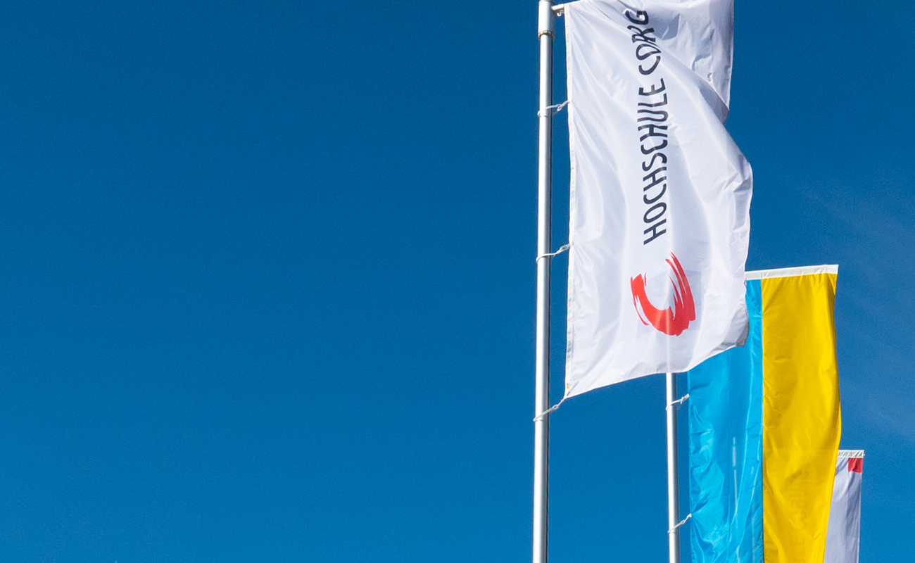 Vor einem klaren blauen Himmel wehen stolz drei Flaggen. Die weiße Flagge im Vordergrund zeigt deutlich „Hochschule Coburg“ neben einem auffälligen roten Logo. Die anderen beiden Flaggen, leicht verdeckt, zeigen leuchtend gelbe und blaue vertikale Streifen, die der Szene einen Farbtupfer verleihen.