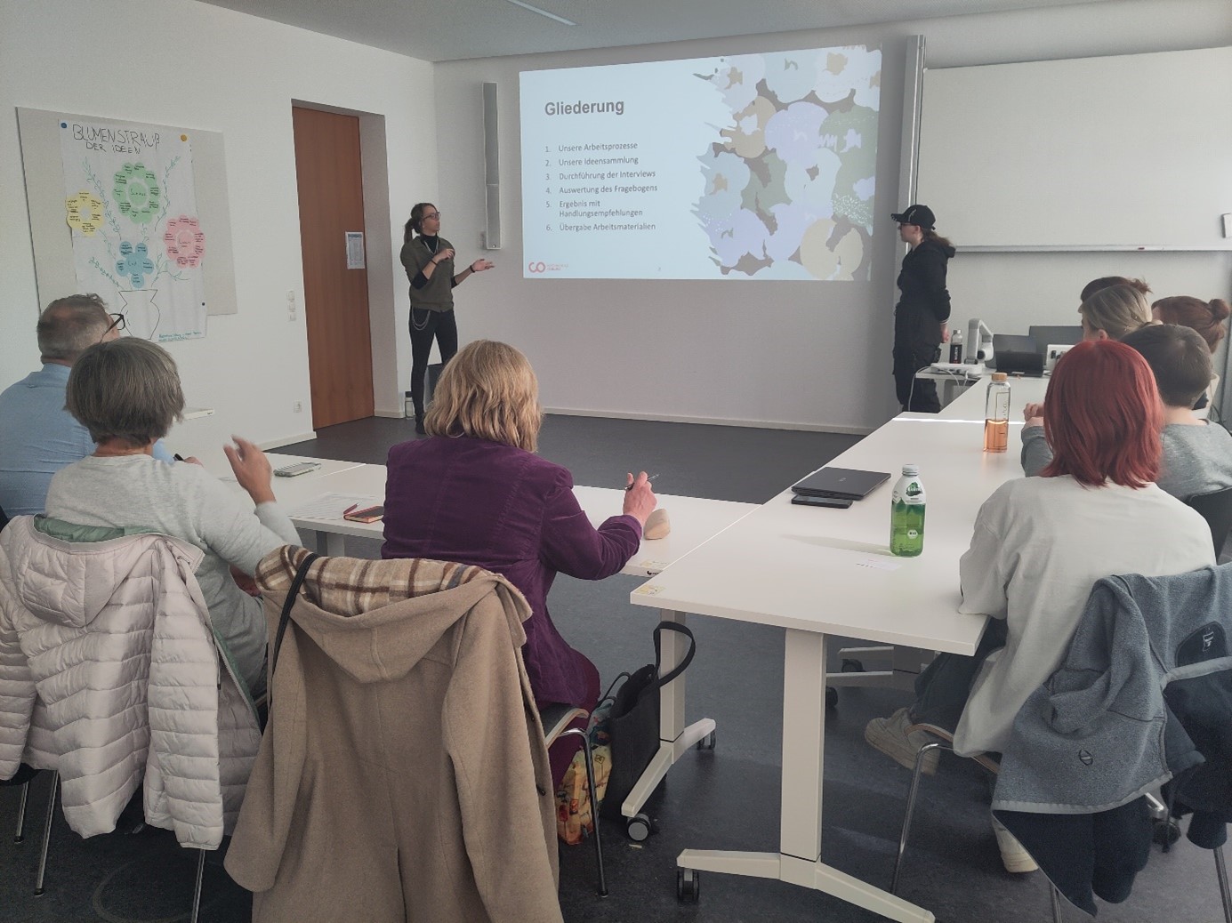 Eine Gruppe von Menschen sitzt an einem Konferenztisch und schaut sich eine Präsentation an. Zwei Vortragende stehen neben einem Bildschirm, auf dem eine Folie mit dem Titel „Gliederung“ läuft, auf der Stichpunkte aufgelistet sind. Beobachter, vermutlich von der Hochschule Coburg, machen sich eifrig Notizen.