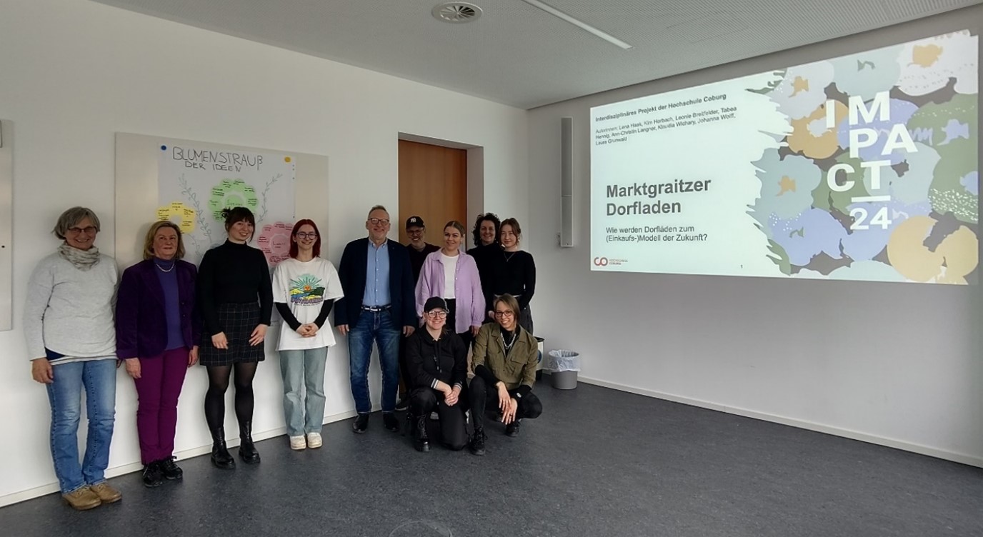 Eine Gruppe von Menschen steht in einem Raum und posiert für ein Foto. Auf der Wand sind die Projektionen „Marktgraitzer Dorfladen“, „Hochschule Coburg“ und „IMPACT 24“ zu sehen. Eine bunte Karte und eine Pinnwand sorgen für Lebendigkeit im Hintergrund.