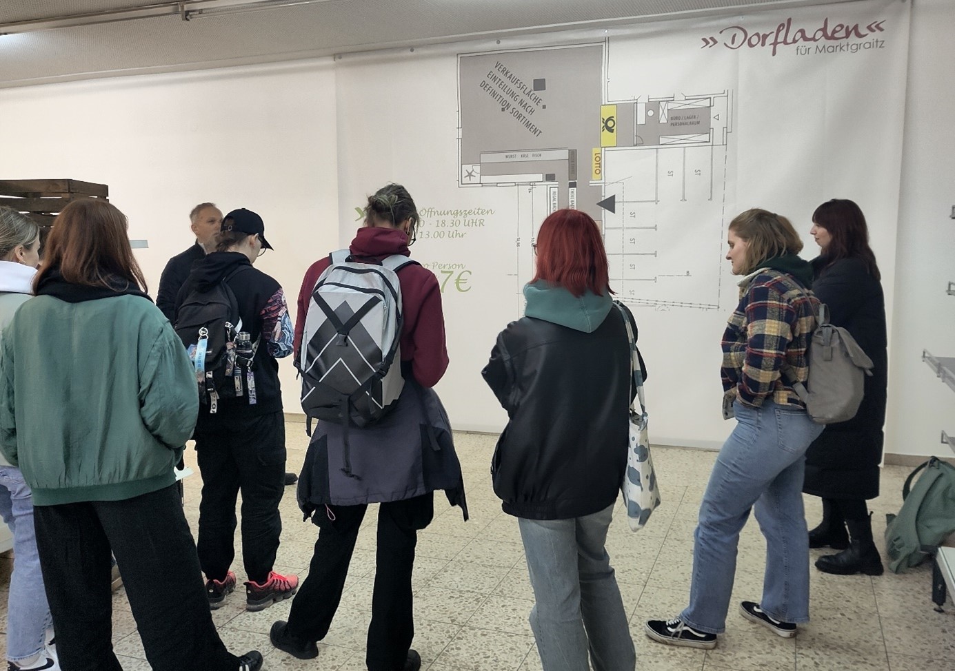 Eine Gruppe von Menschen steht in einem Raum der Hochschule Coburg vor einer Wand mit Bauplänen. Die meisten tragen Rucksäcke und sind leger gekleidet. Der Raum hat einen gefliesten Boden und Neonlicht, was eine Atmosphäre schafft, die zum Lernen und Zusammenarbeiten einlädt.