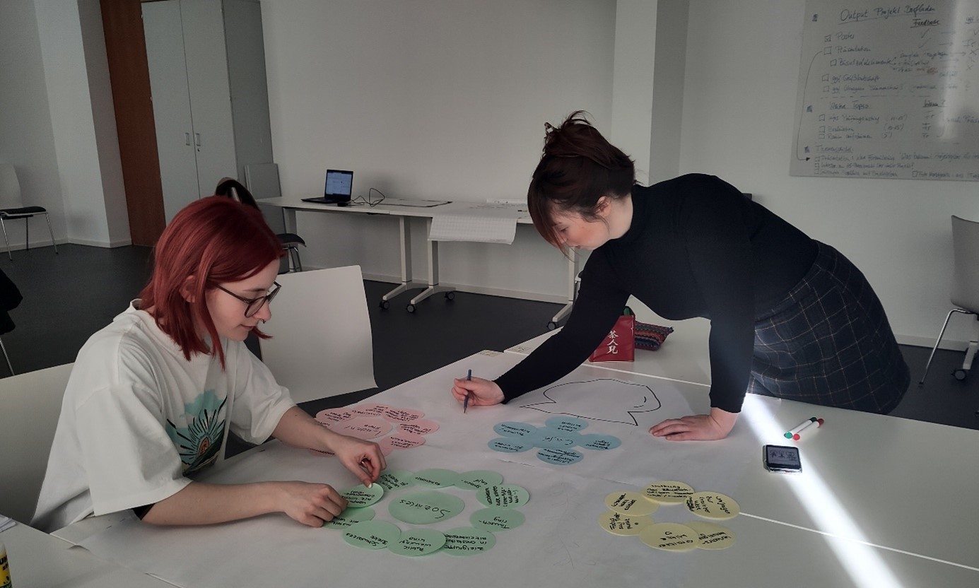 Two people are collaborating in a bright room at Hochschule Coburg, arranging colorful paper shapes and jotting down notes on a large sheet spread across the table. A laptop hums quietly in the background, while a whiteboard on the wall displays visible text.