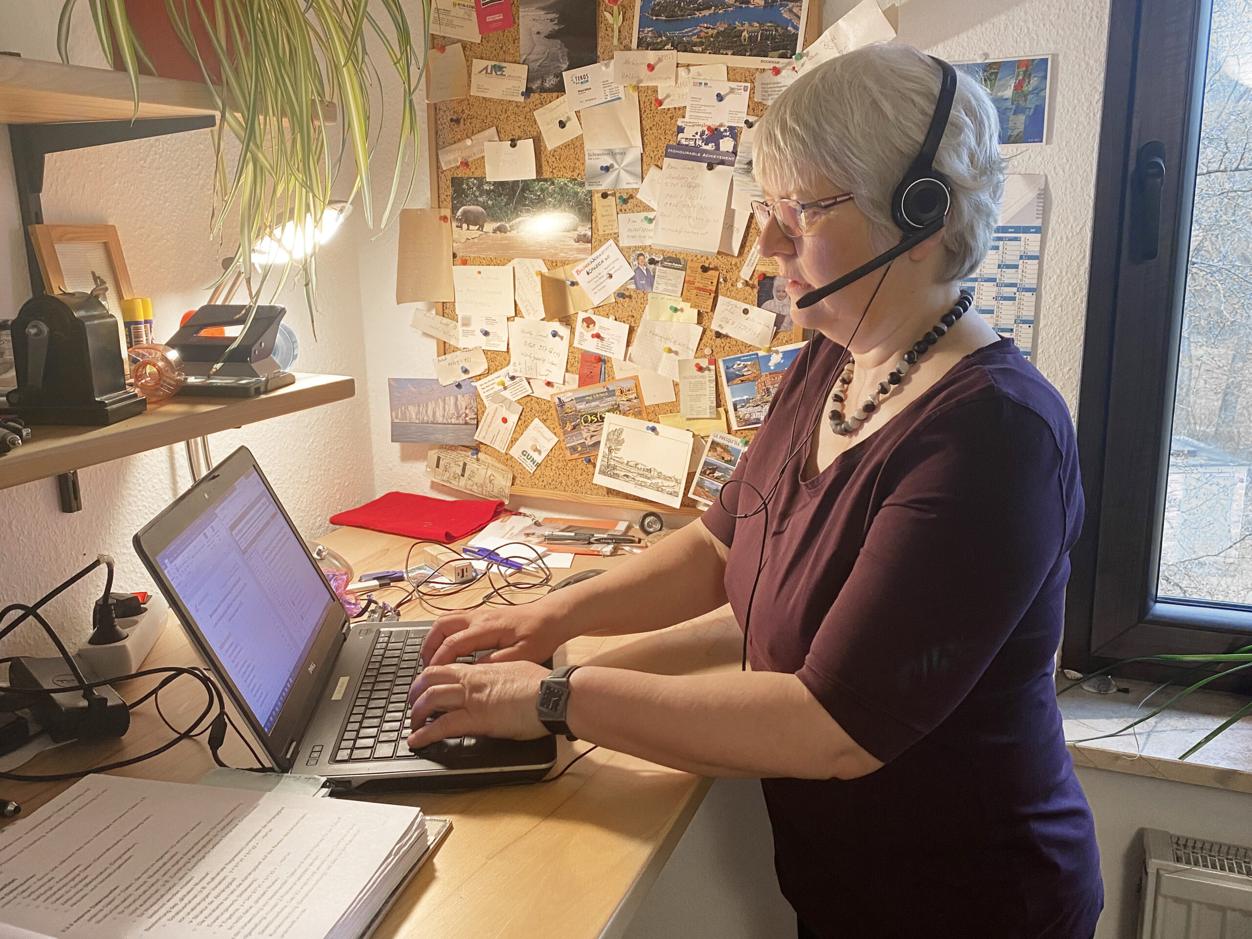 Eine ältere Frau steht mit Headset an einem überladenen Schreibtisch und tippt auf ihrem Laptop. In dem gut beleuchteten Raum, der mit Erinnerungsstücken der Hochschule Coburg geschmückt ist, ist ein schwarzes Brett mit Postkarten und Notizen bedeckt. Eine Pflanze hängt von einem Regal über dem Schreibtisch und verleiht ihrer akademischen Oase einen Hauch von Natur.