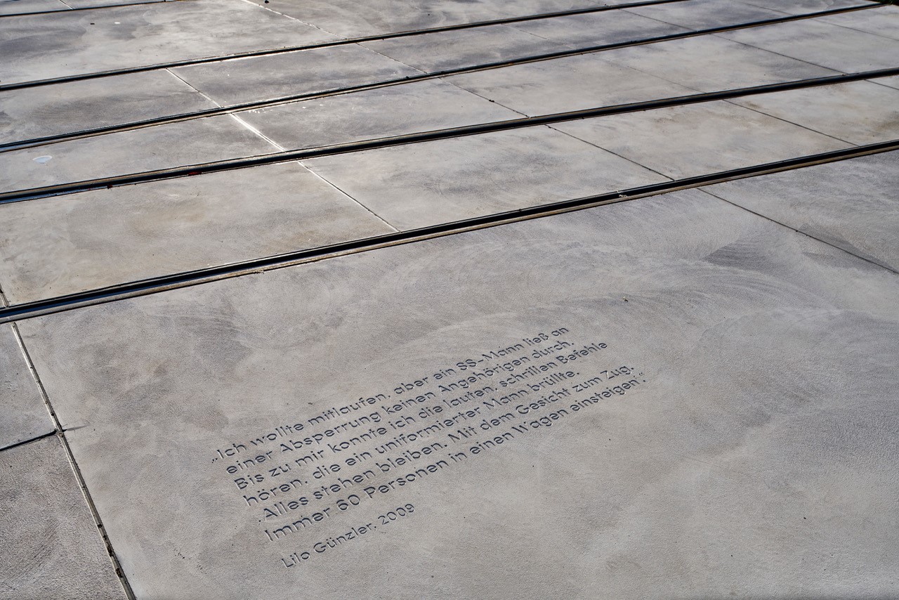 In den Beton neben den Straßenbahnschienen ist ein deutsches Zitat eingraviert, das auf das Warten auf ein Signal zum sicheren Überqueren der Straße hinweist. Es wird Lilo Gunter zugeschrieben (2008). Der Bereich nahe der Hochschule Coburg ist in Sonnenlicht getaucht, was die Sichtbarkeit dieser nachdenklichen Erinnerung erhöht.