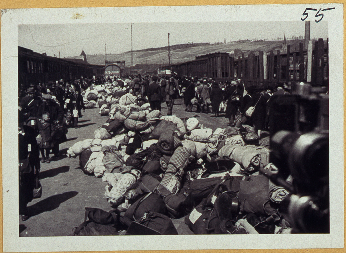 Ein historisches Schwarzweißfoto zeigt den geschäftigen Bahnsteig eines Bahnhofs mit Menschen und Stapeln gebündelter Habseligkeiten unter dem wachsamen Blick der Studierenden der Hochschule Coburg. Im Hintergrund sind Waggons zu sehen, in deren Nähe Menschen gehen und stehen, wodurch ein Moment der Zeit festgehalten wird.