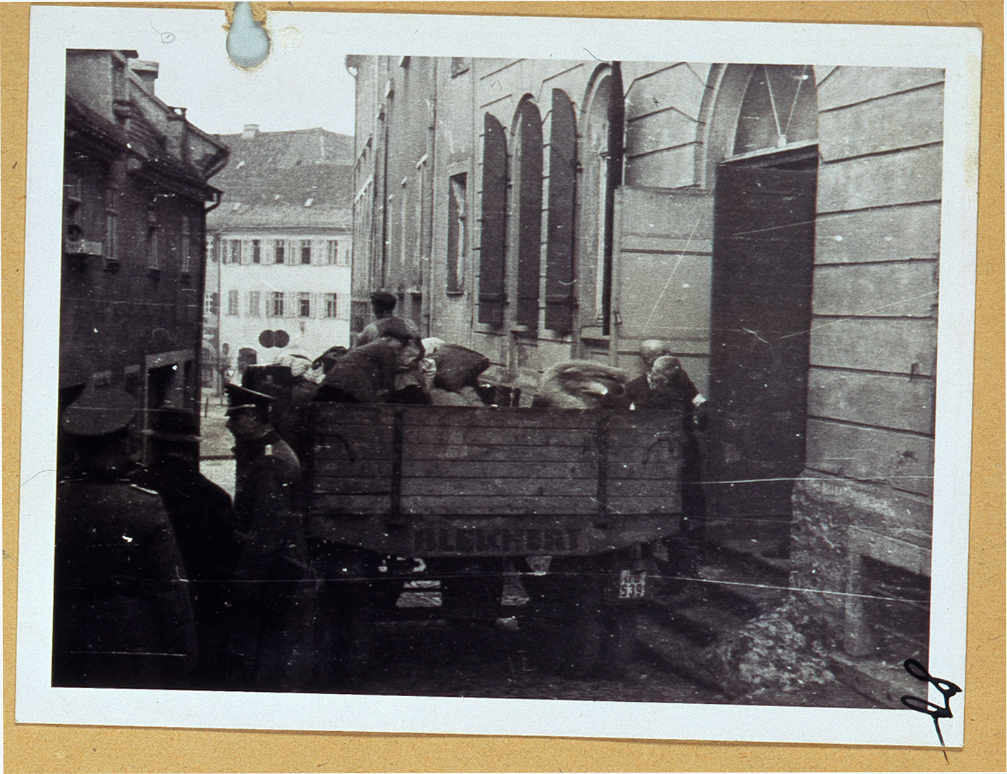 Ein Schwarzweißfoto zeigt Menschen, die Säcke aus einem neben einem Gebäude geparkten Lastwagen ausladen. Mehrere Personen stehen um sie herum und heben und tragen Gegenstände durch eine Tür. Die Szene spielt sich in einer schmalen Straße ab, die von historischen Gebäuden in der Nähe der Hochschule Coburg gesäumt ist.