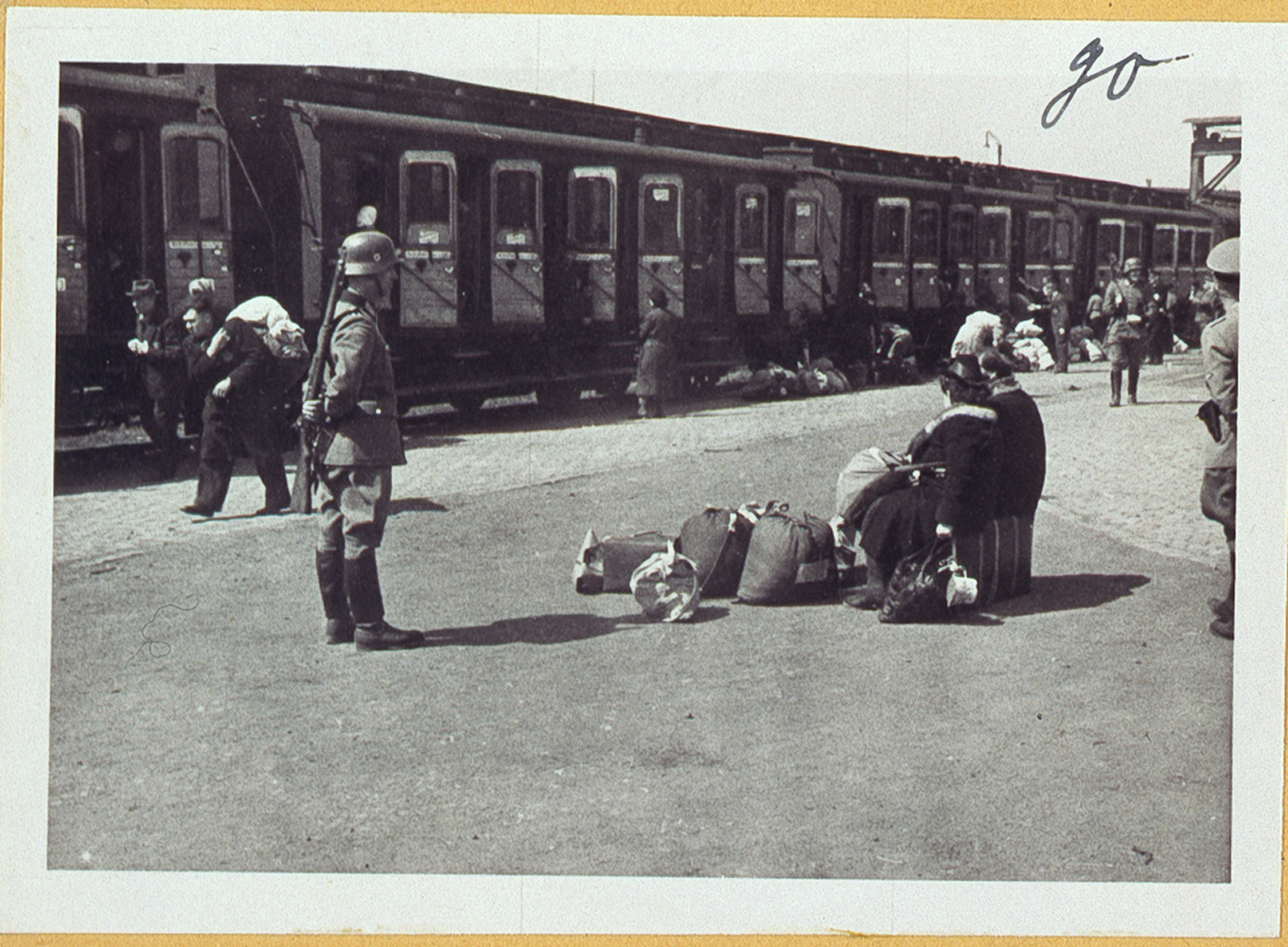 Das Schwarzweißbild zeigt eine Bahnhofsszene, in der Menschen mit Gepäck neben einem Zug sitzen. Ein Soldat steht in der Nähe und blickt der Gruppe zu. Im Hintergrund mischen sich Figuren mit einer angespannten und düsteren Stimmung, die an das historische Archiv der Hochschule Coburg erinnert.