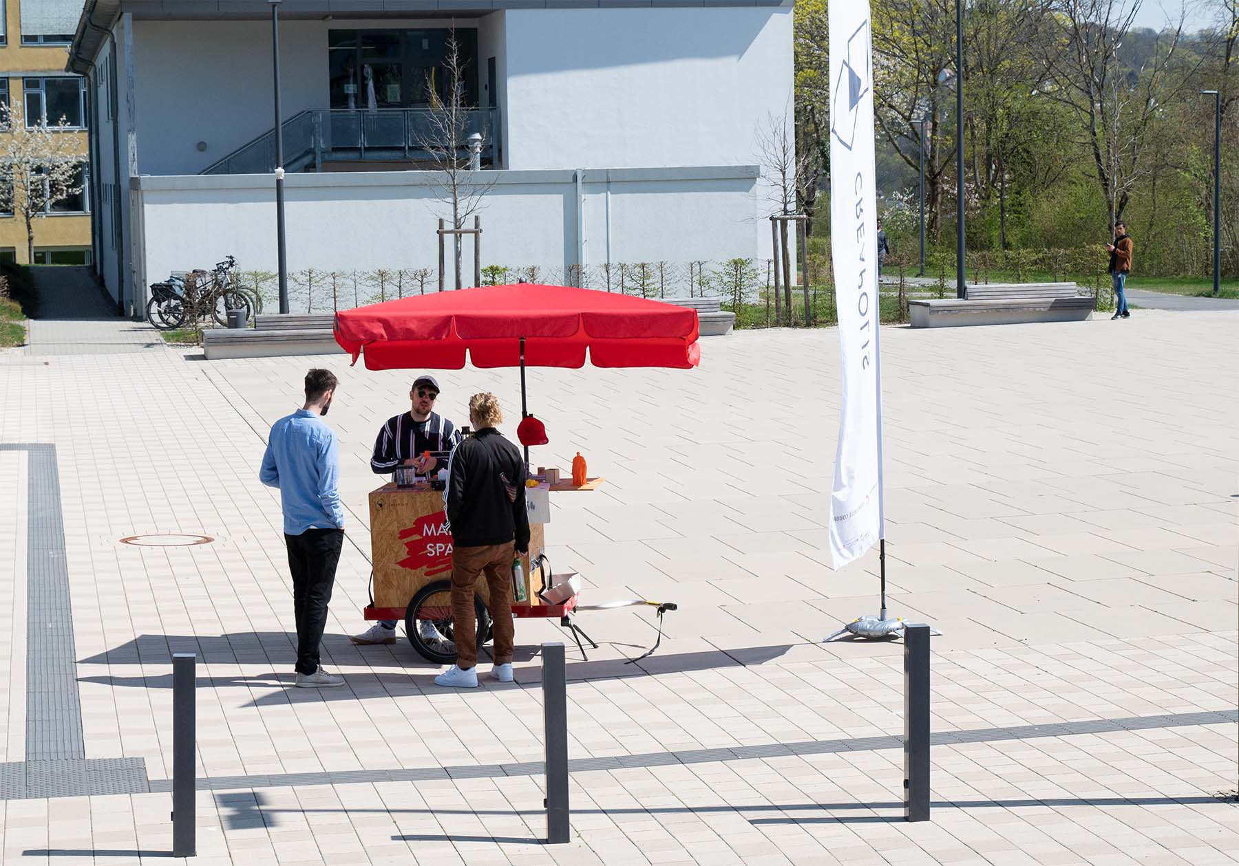 Zwei Menschen stehen an einem kleinen Wagen mit rotem Dach, an dem ein Verkäufer der Hochschule Coburg ihnen Getränke serviert. Auf dem Wagen steht "Make Space". Rechts flattert eine weiße Fahne mit einem Logo auf dem hellen, sonnenbeschienenen Platz.