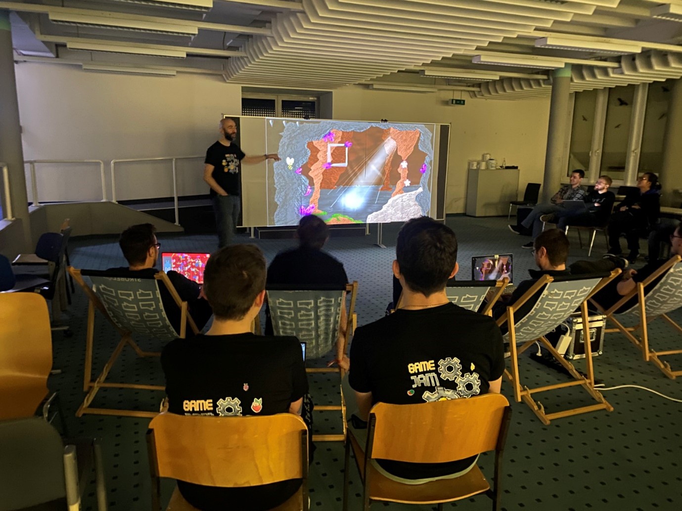 In a dimly lit room at Hochschule Coburg, a group of people sits on chairs and loungers, captivated by a colorful video game presentation projected on the screen. The presenter stands nearby, bringing the vibrant world to life while engaging with the curious audience.