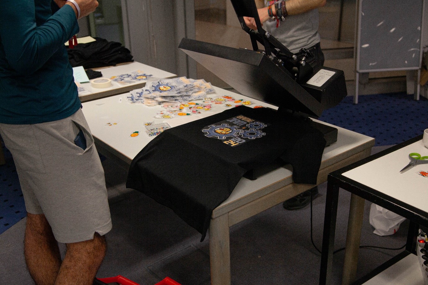 A person is operating a heat press machine, pressing designs onto a black T-shirt, perhaps for a Hochschule Coburg event. The table is cluttered with various cut-out designs as another person in a teal shirt and gray shorts assists. Scissors and small tools are visible amidst the creative chaos.