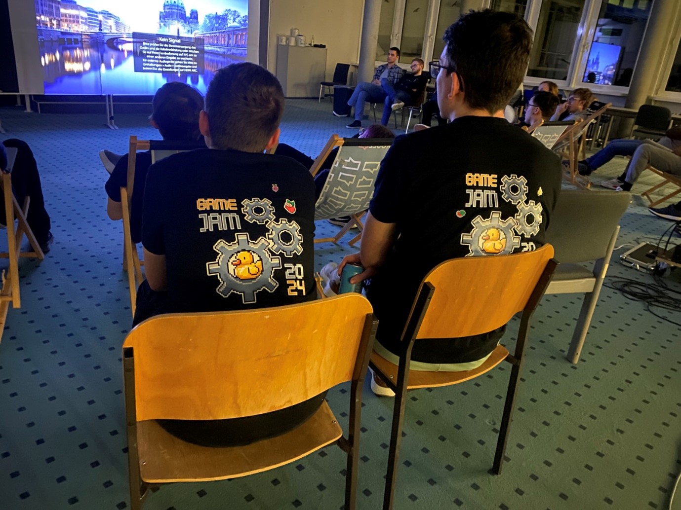 Two people sit on wooden chairs at a Hochschule Coburg event, wearing matching "Game Jam 2024" T-shirts adorned with gear and cartoon character designs. A large screen at the front displays event information in the dimly lit room, with more attendees seated in the background.