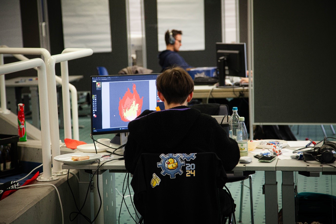 A person sits at a desk at Hochschule Coburg, with a computer displaying a fiery image. The setting appears to be an office or classroom with others working busily. Snacks and a drink are on the table, and the individual wears a black hoodie adorned with a 2024 logo.