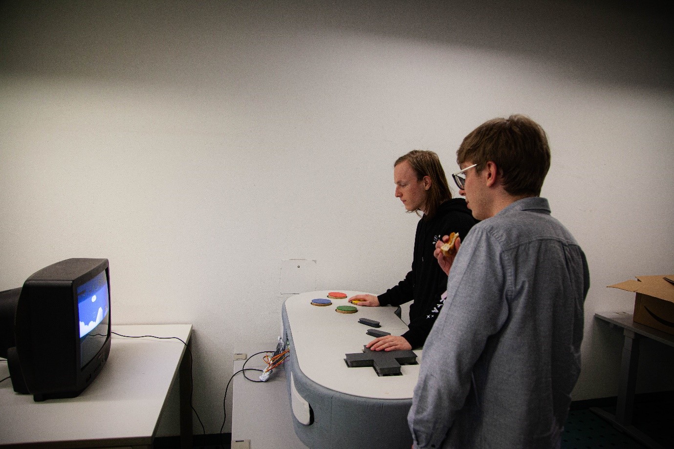 Two people stand at a gaming console with colorful buttons, interacting with a retro TV screen displaying a video game. One person holds a snack while looking at the screen. The room, reminiscent of Hochschule Coburg’s relaxed vibe, is simple and appears to be a casual setting.