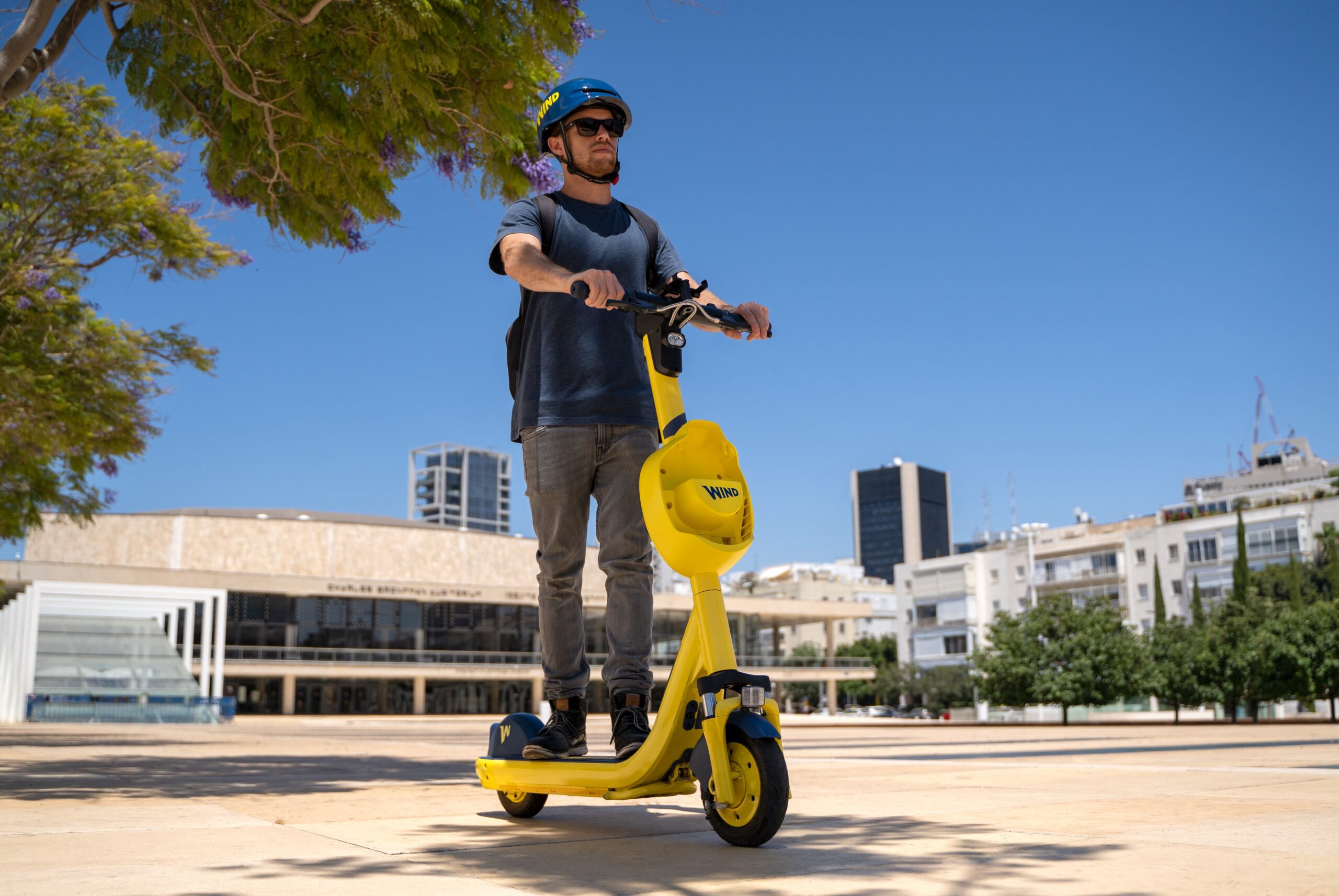 Bei klarem Himmel rast eine Person mit Sonnenbrille und Helm auf einem leuchtend gelben Elektroroller über einen Stadtplatz. Die Kulisse aus modernen Gebäuden und Bäumen lässt den lebendigen Geist der Hochschule Coburg erahnen.