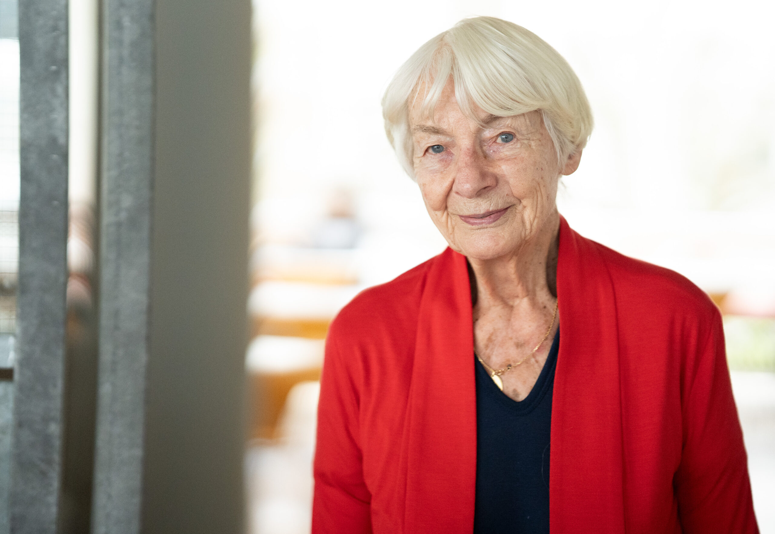 An elderly woman with short white hair stands indoors at Hochschule Coburg, wearing a red cardigan over a black top. She is smiling gently, with a neutral-toned background that appears softly blurred.