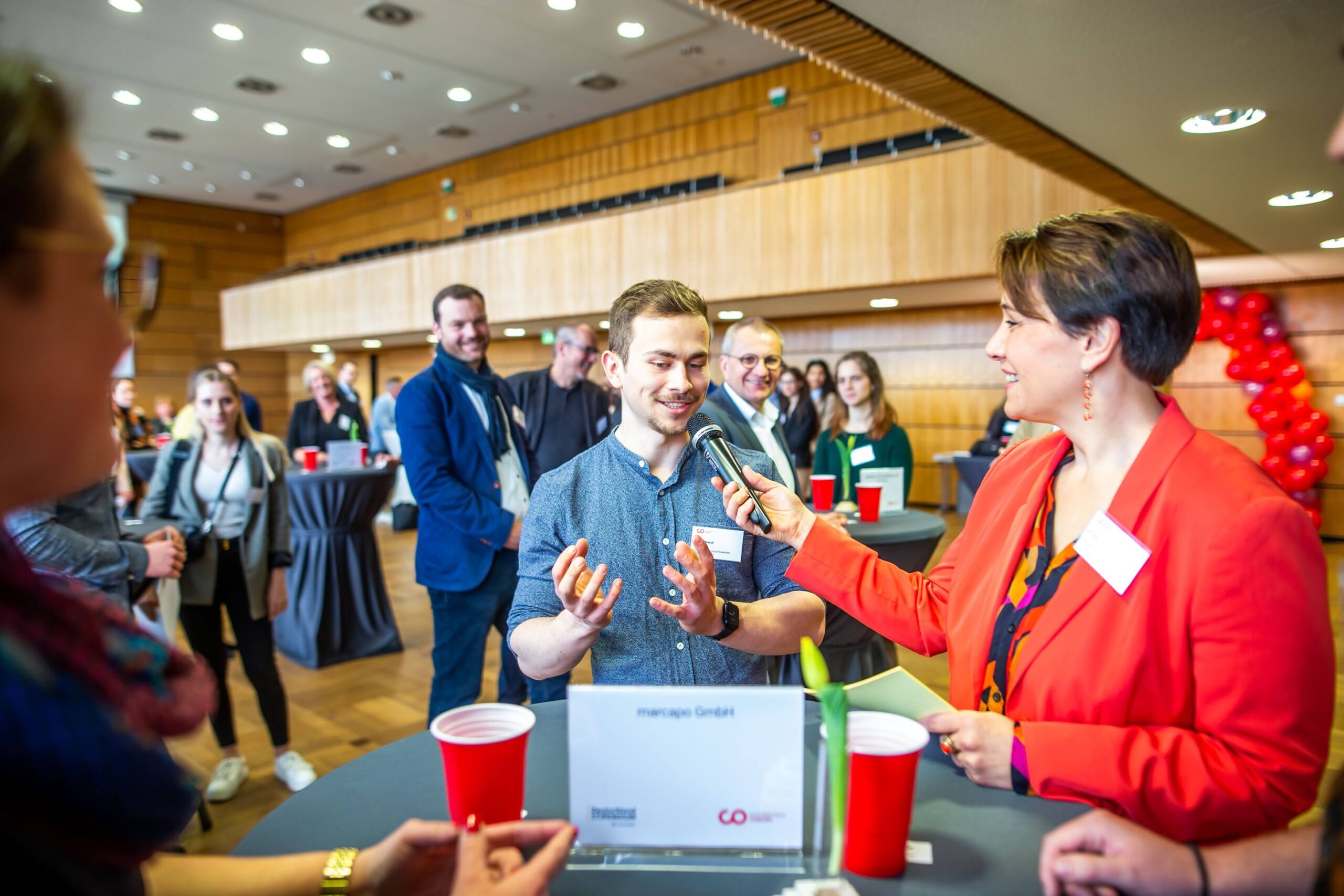 Bei einer Networking-Veranstaltung der Hochschule Coburg in einem Konferenzraum hält eine Frau in einem roten Blazer ein Mikrofon und unterhält sich mit einem Mann, der enthusiastisch gestikuliert. Die Teilnehmer sitzen bei Getränken an Tischen und rote und schwarze Luftballons sorgen für eine lebhafte Atmosphäre.