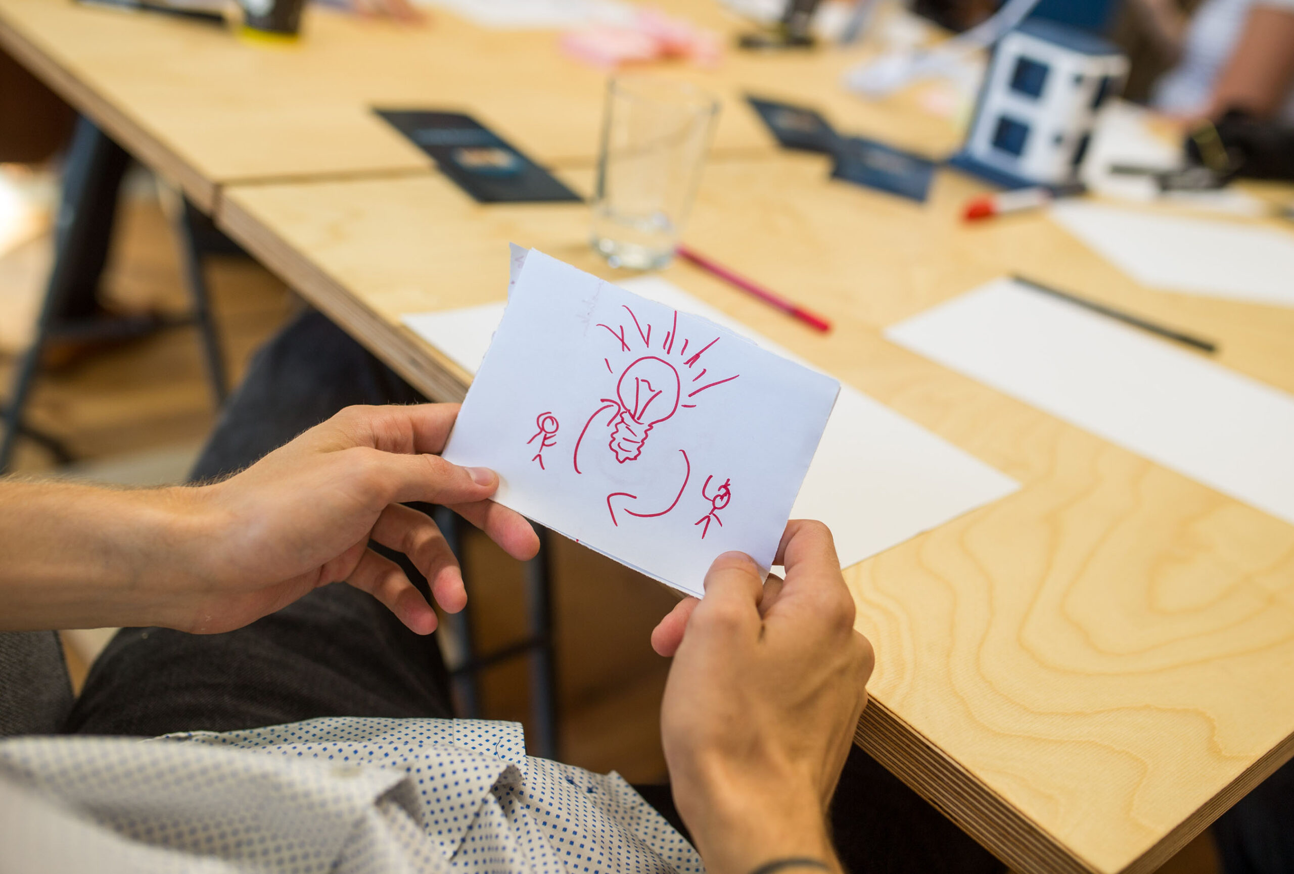 Eine Person hält ein weißes Blatt Papier mit der roten Zeichnung einer Glühbirne und zwei Strichmännchen in der Hand, die die Kreativität an der Hochschule Coburg symbolisieren. Im Hintergrund ist ein Holztisch zu sehen, auf dem verstreute Papiere, ein Stift und ein Glas herumliegen.