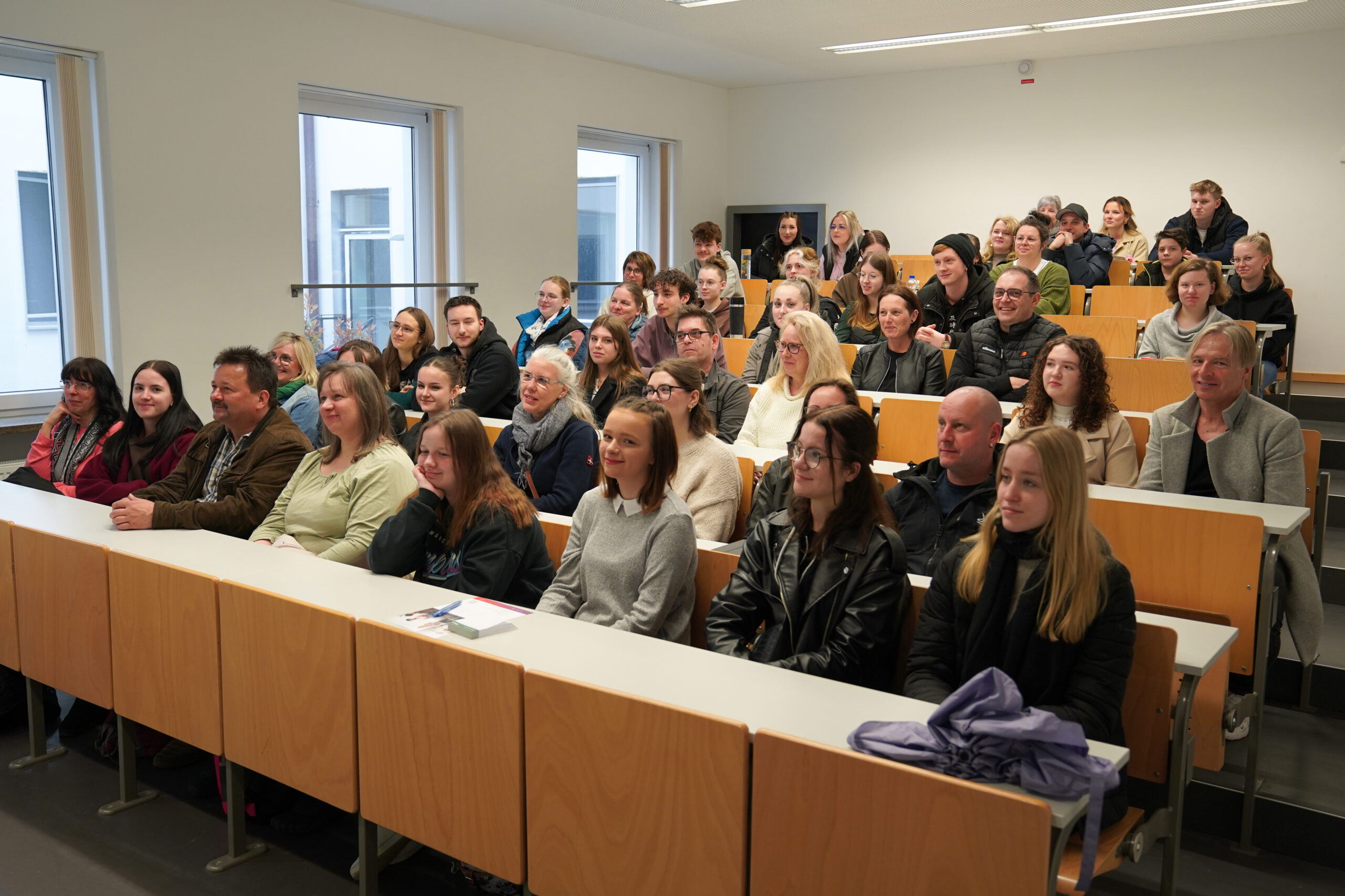 A diverse group of people sits attentively in a lecture hall at Hochschule Coburg, occupying several rows of wooden desks. The room is well-lit with natural light streaming through large windows on the side.