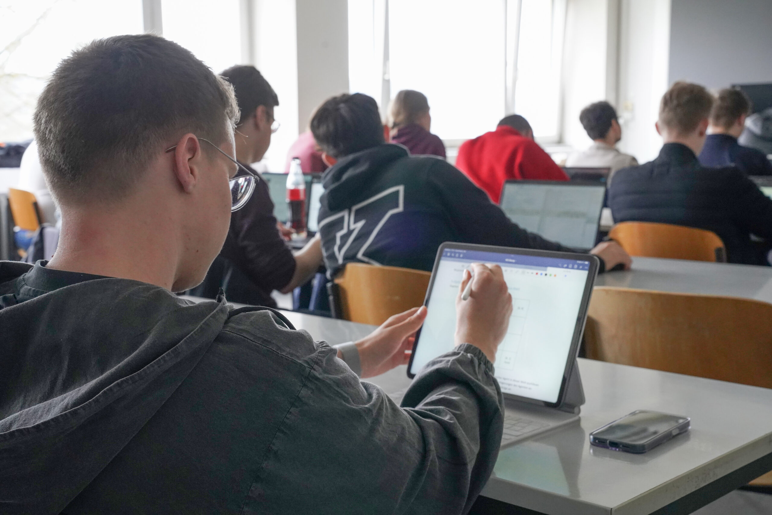 In a bright, naturally lit classroom at Hochschule Coburg, a student writes on a tablet using a stylus. Seated at a desk with a small pouch nearby, they are surrounded by classmates equally focused on their work, all benefiting from the serene atmosphere created by the light streaming through large windows.