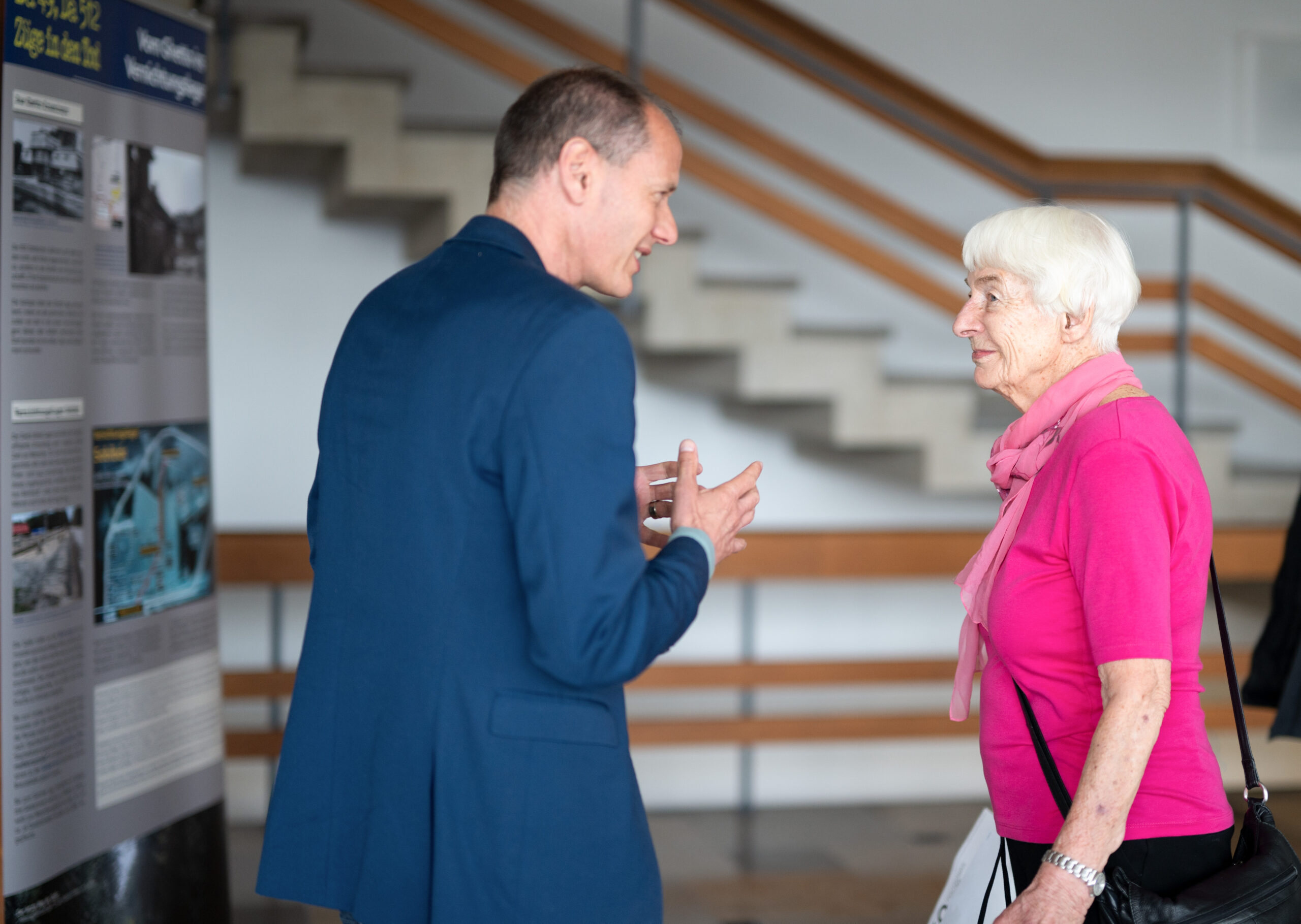 Ein Mann im blauen Anzug und eine ältere Frau in rosa Oberteil und Schal unterhalten sich an der Hochschule Coburg. Sie befinden sich in einem Innenraum mit Informationsplakaten und einer Treppe im Hintergrund.